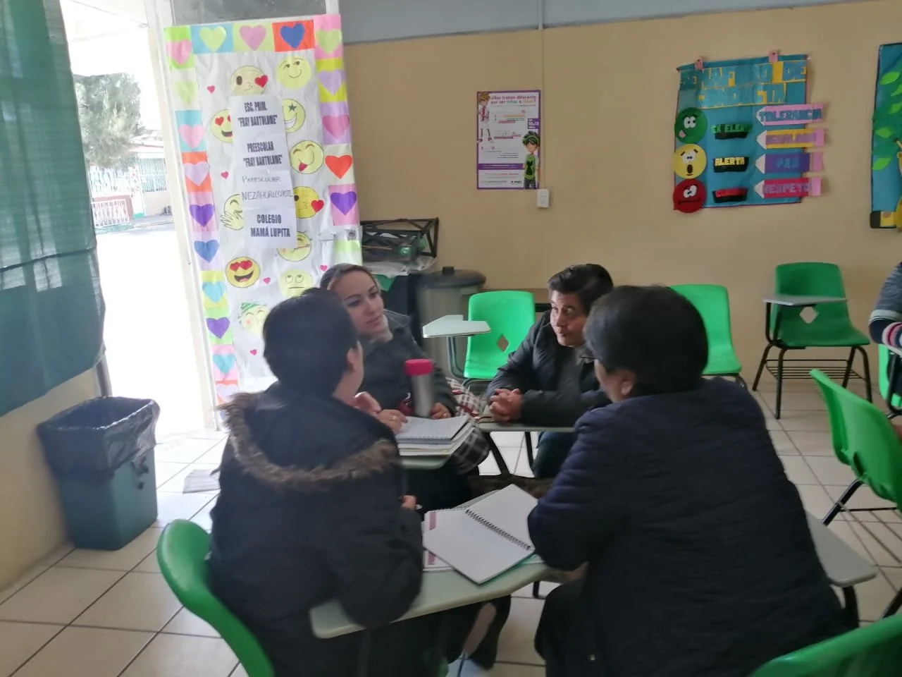 Cuatro mujeres conversando en una mesa en una sala escolar decorada con carteles y retratos coloridos.