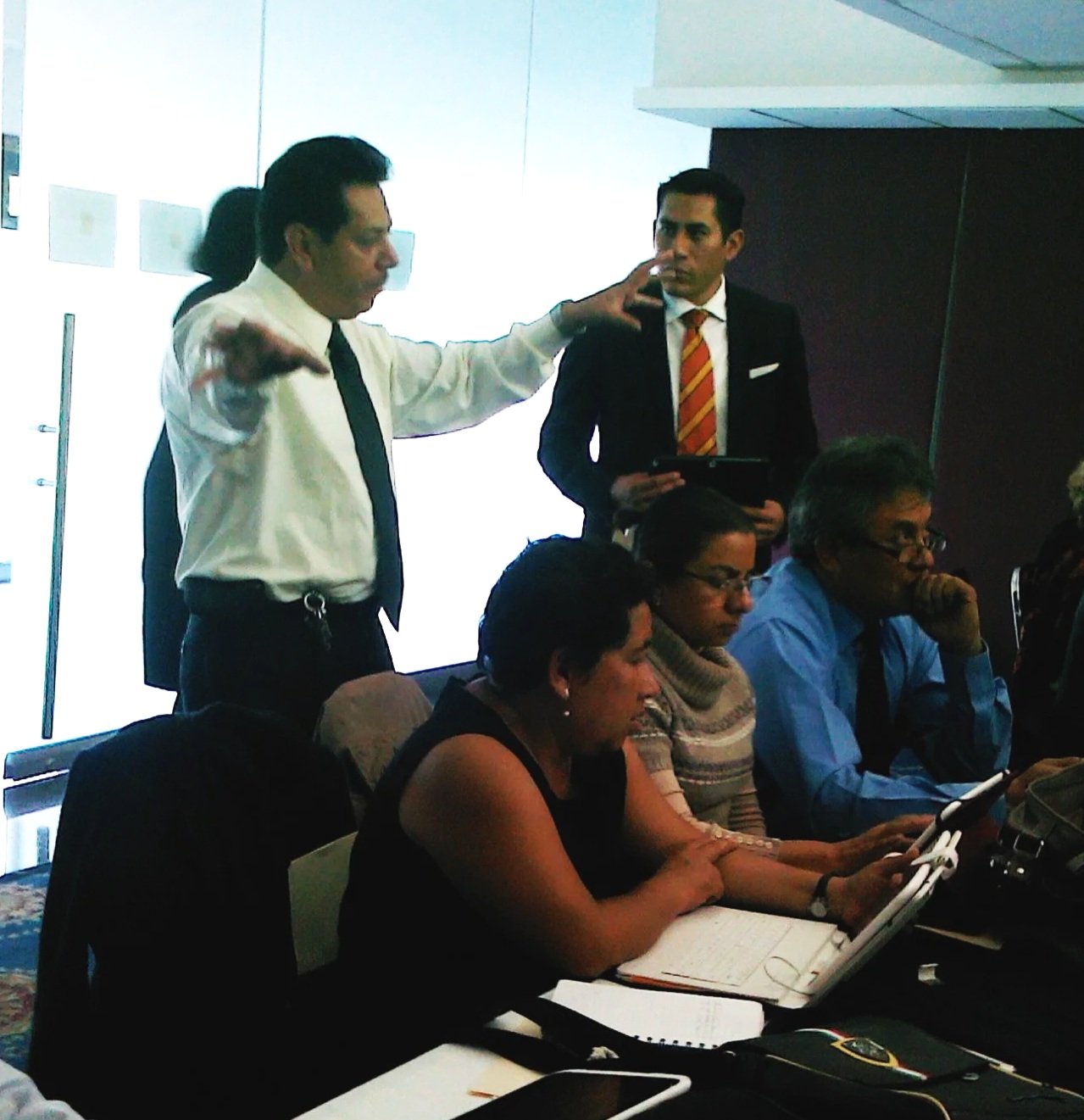 Reunión de oficina con varios asistentes, algunos tomando notas o usando dispositivos electrónicos, y una persona de pie que parece estar dando una explicación o discusión.