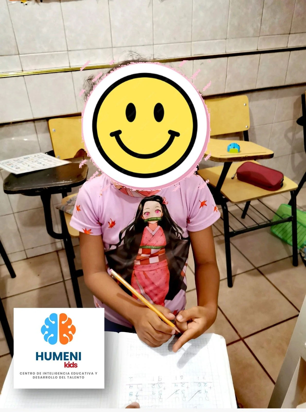Niña en aula con rostro cubierto con cara sonriente, usando camiseta con personaje anime, escribiendo en cuaderno. Fondo con sillas, objetos y paredes de azulejos. Logo de HUMENI Kids en esquina.