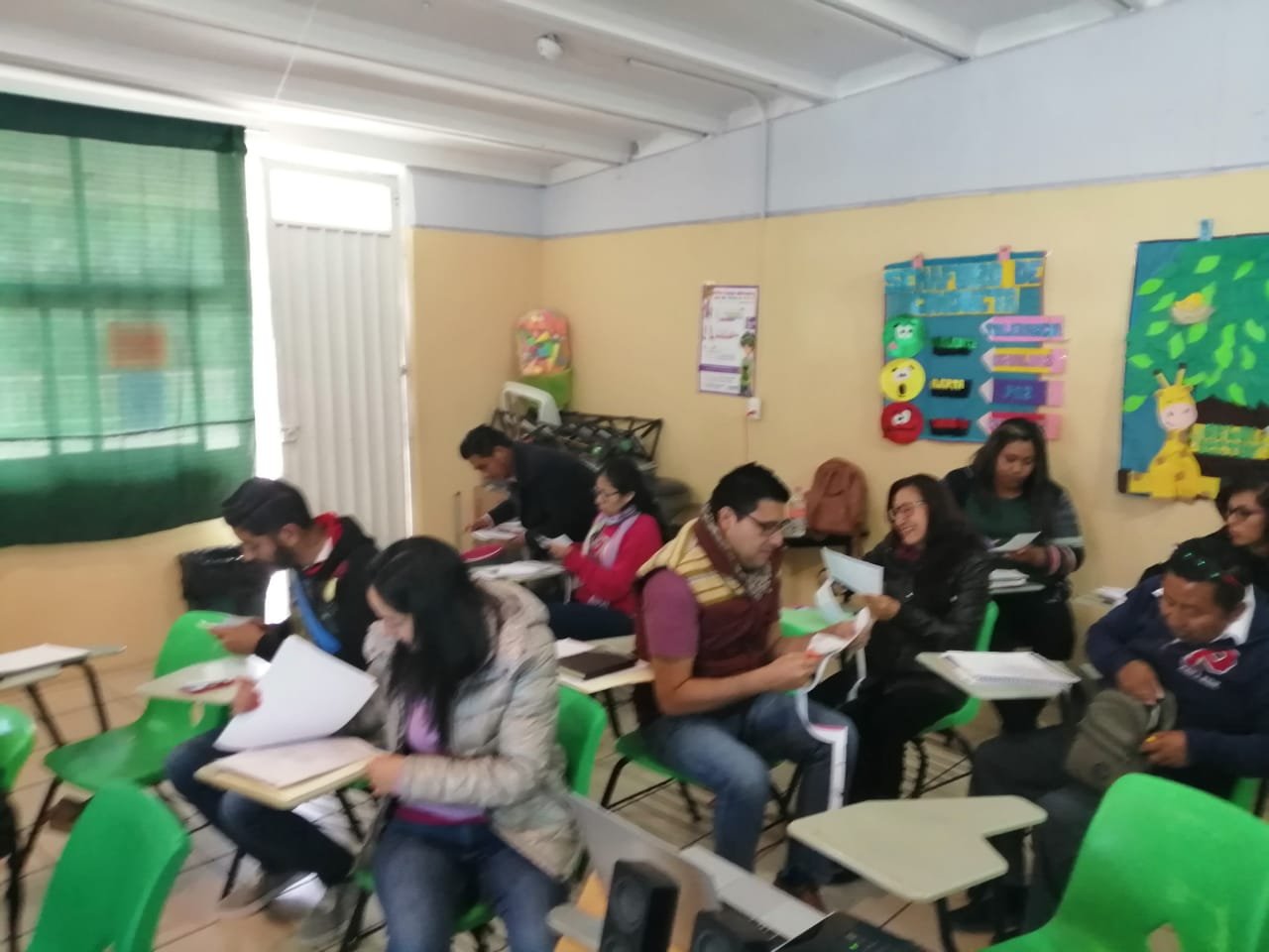 Grupo de adultos en un aula, leyendo libros y documentos, con paredes decoradas con dibujos y materiales educativos.