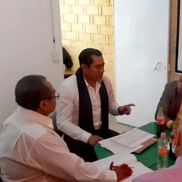 Hombres en una reunión, uno de ellos hablando con documentos en la mesa y botellas de bebidas