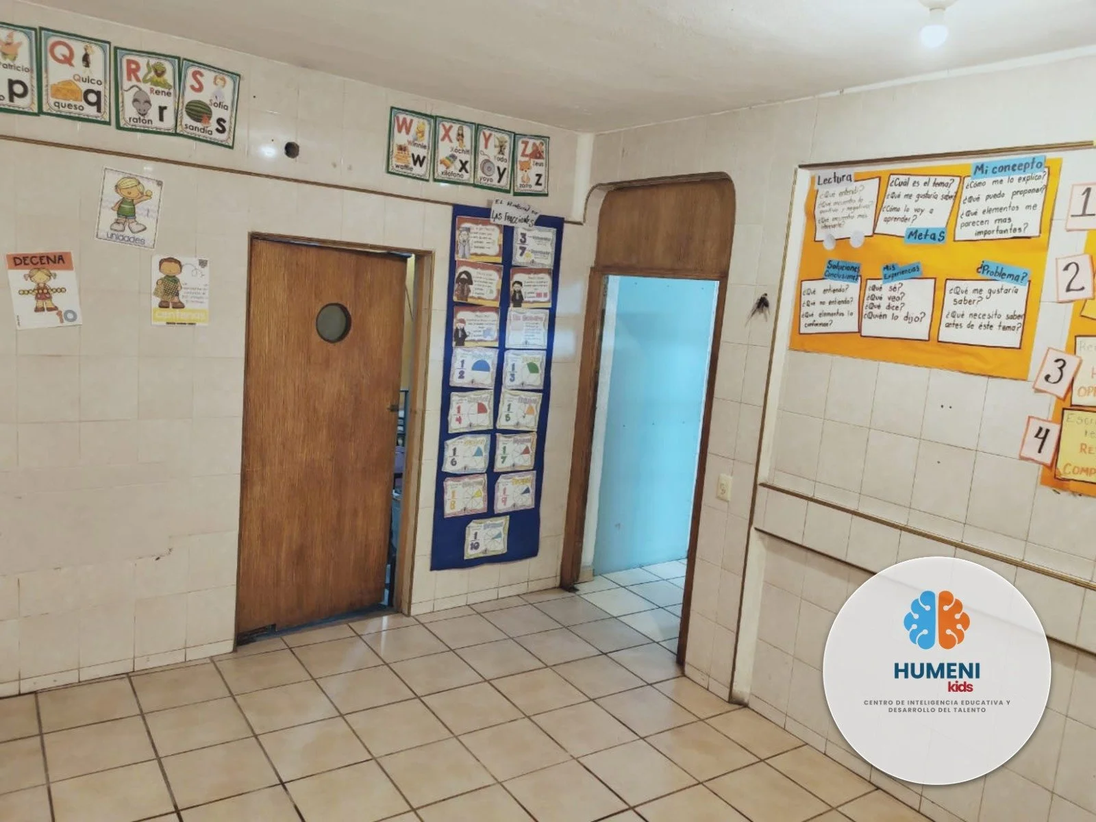 Aula escolar con paredes de azulejos blancas, con carteles y posters educativos, incluyendo procesos de lectura y conceptos matemáticos para niños, puertas de madera y un gráfico en la esquina inferior derecha que dice 'HUMENI kids'.