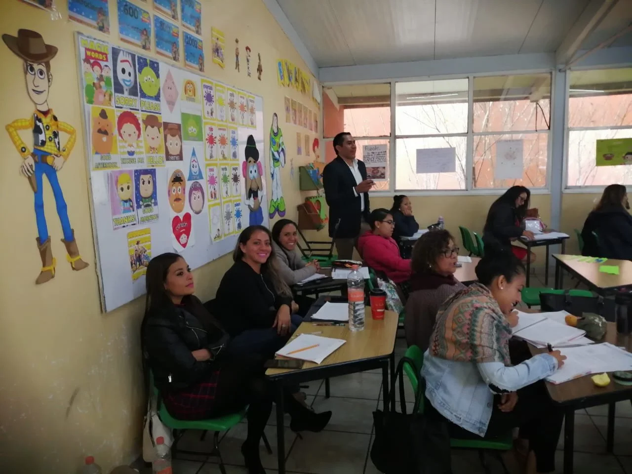 Clase en un aula con varias personas sentadas en mesas, un hombre de pie hablando, y decoraciones de personajes de Toy Story en la pared.
