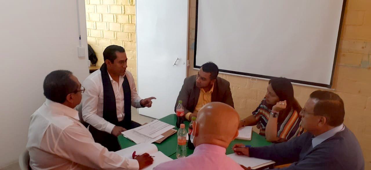 Grupo de seis personas en una reunión en una sala, con documentos, agua y refrescos en la mesa.