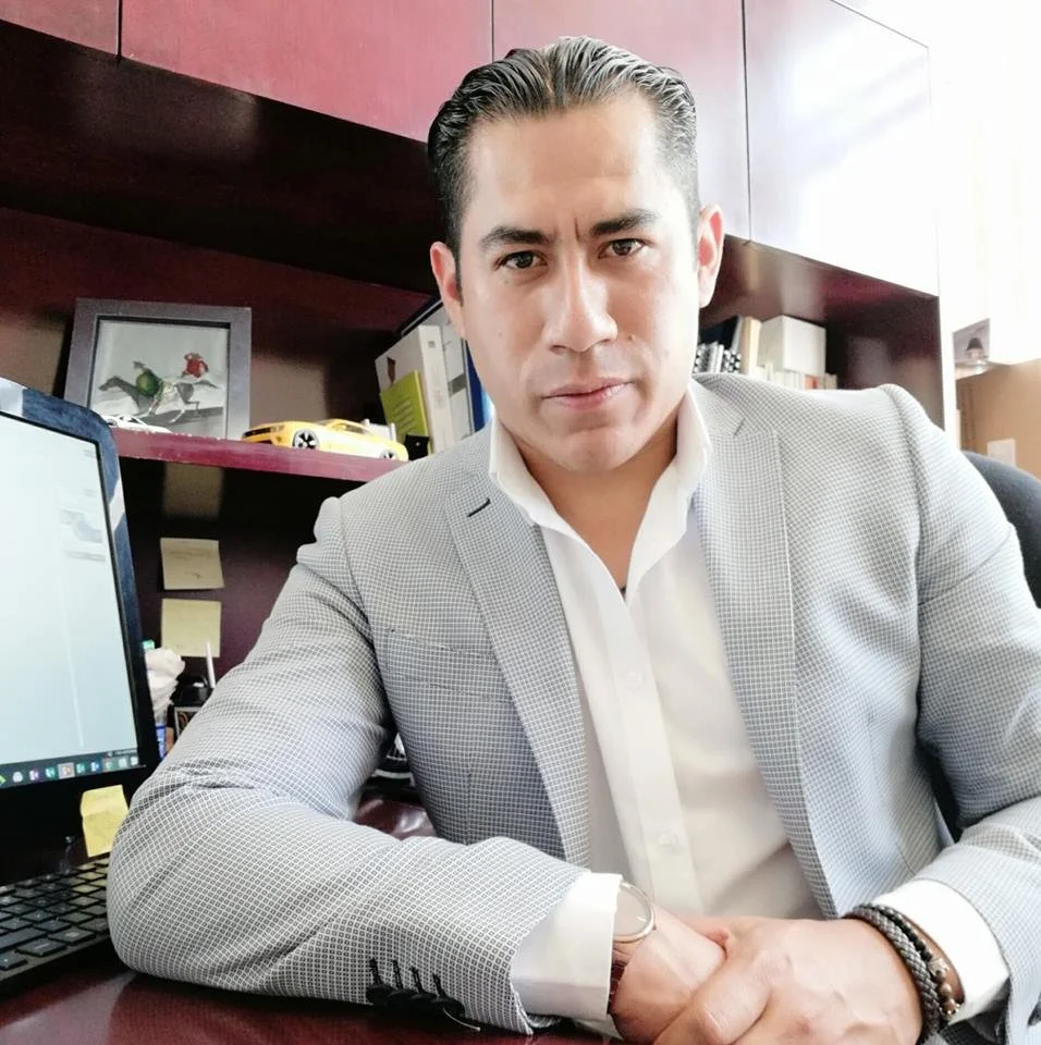 Hombre sentado en oficina con computadora, libros y decoración en el fondo, vestido con saco gris y camisa blanca.