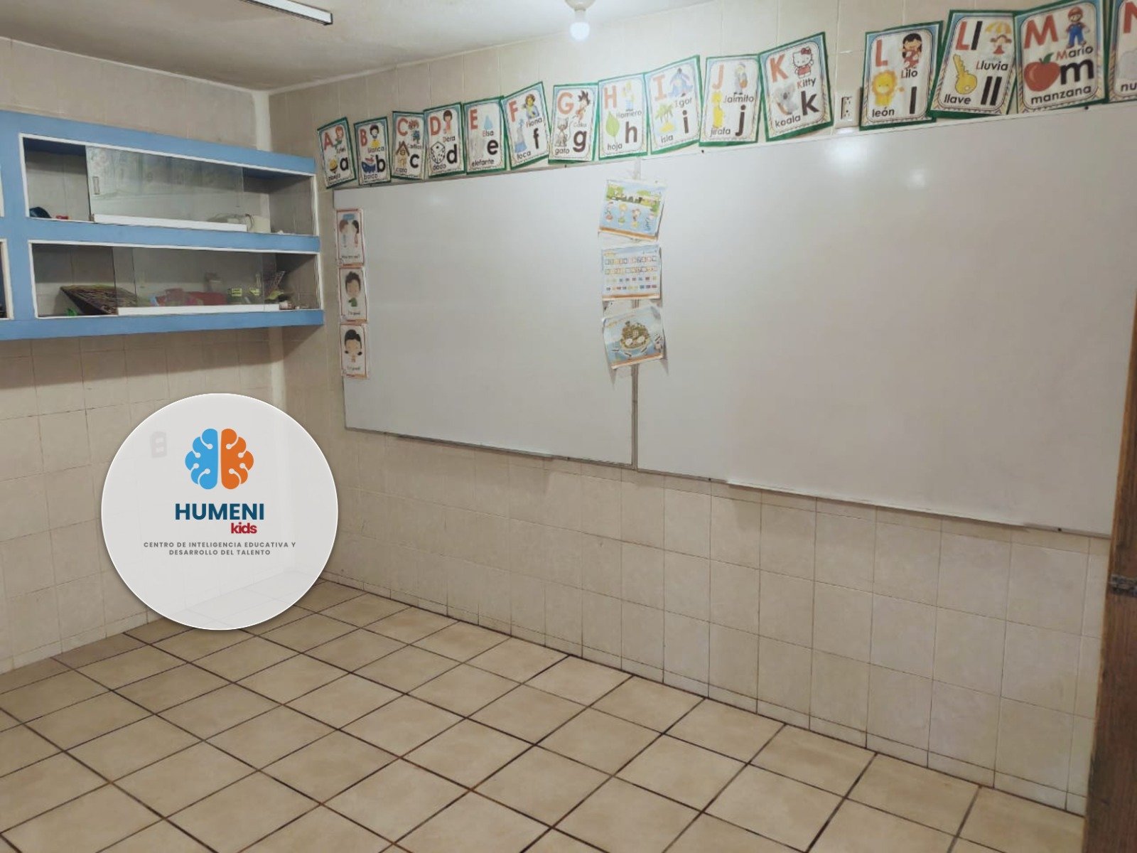 Aula escolar con cartel de letras del alfabeto, dibujos y palabras en español, y cartel de logo de HUMENI Kids que tiene un cerebro dividido en dos colores, azul y naranja.