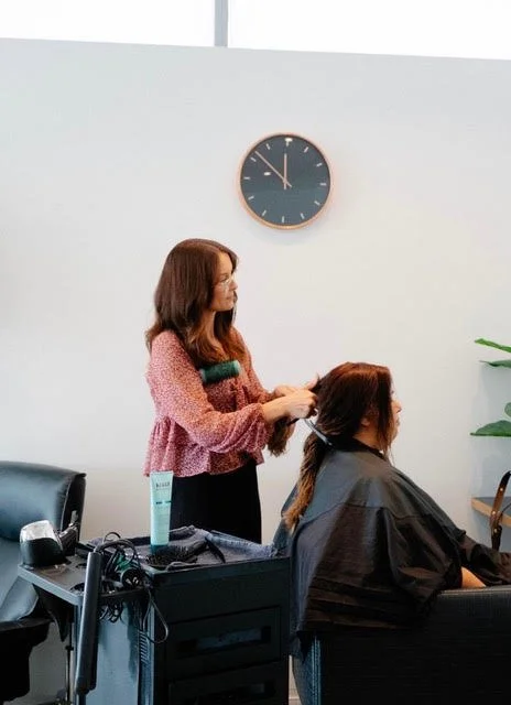 A woman getting her hair styled at a salon with a clock on the white wall behind her.