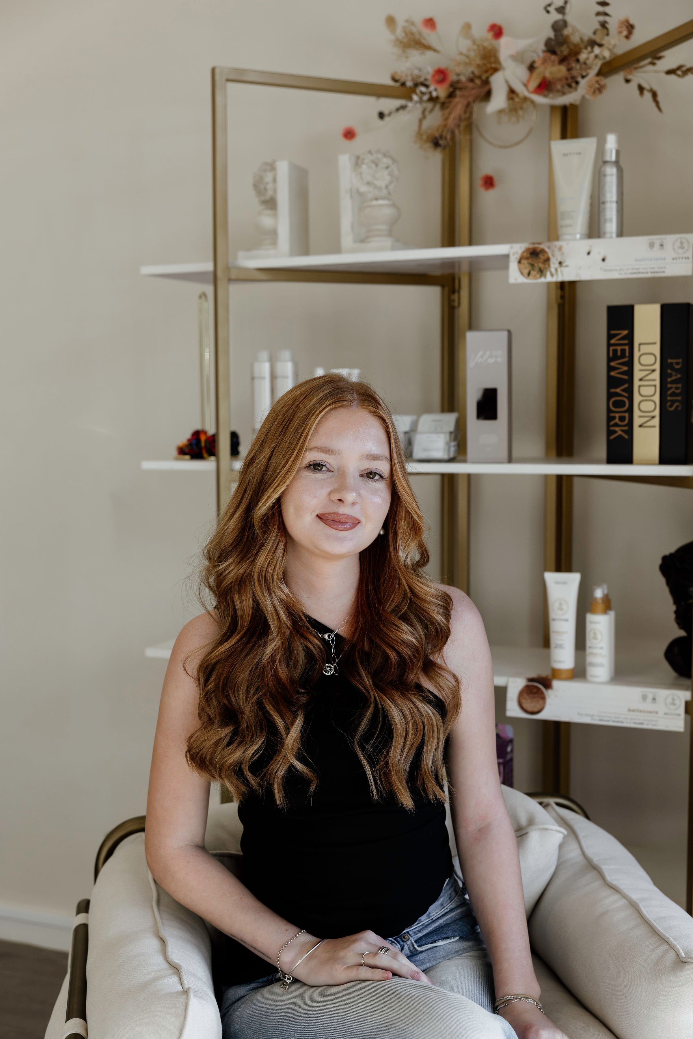 A young woman with long, wavy red hair is sitting on a cream-colored sofa. She has a slight smile, and is wearing a black sleeveless top, several bracelets, and rings. Behind her is a white shelf with decorative items, including dried flowers in vases, books with titles like 'Paris', 'London', and 'New York', as well as skincare products. The background wall is plain and light-colored.