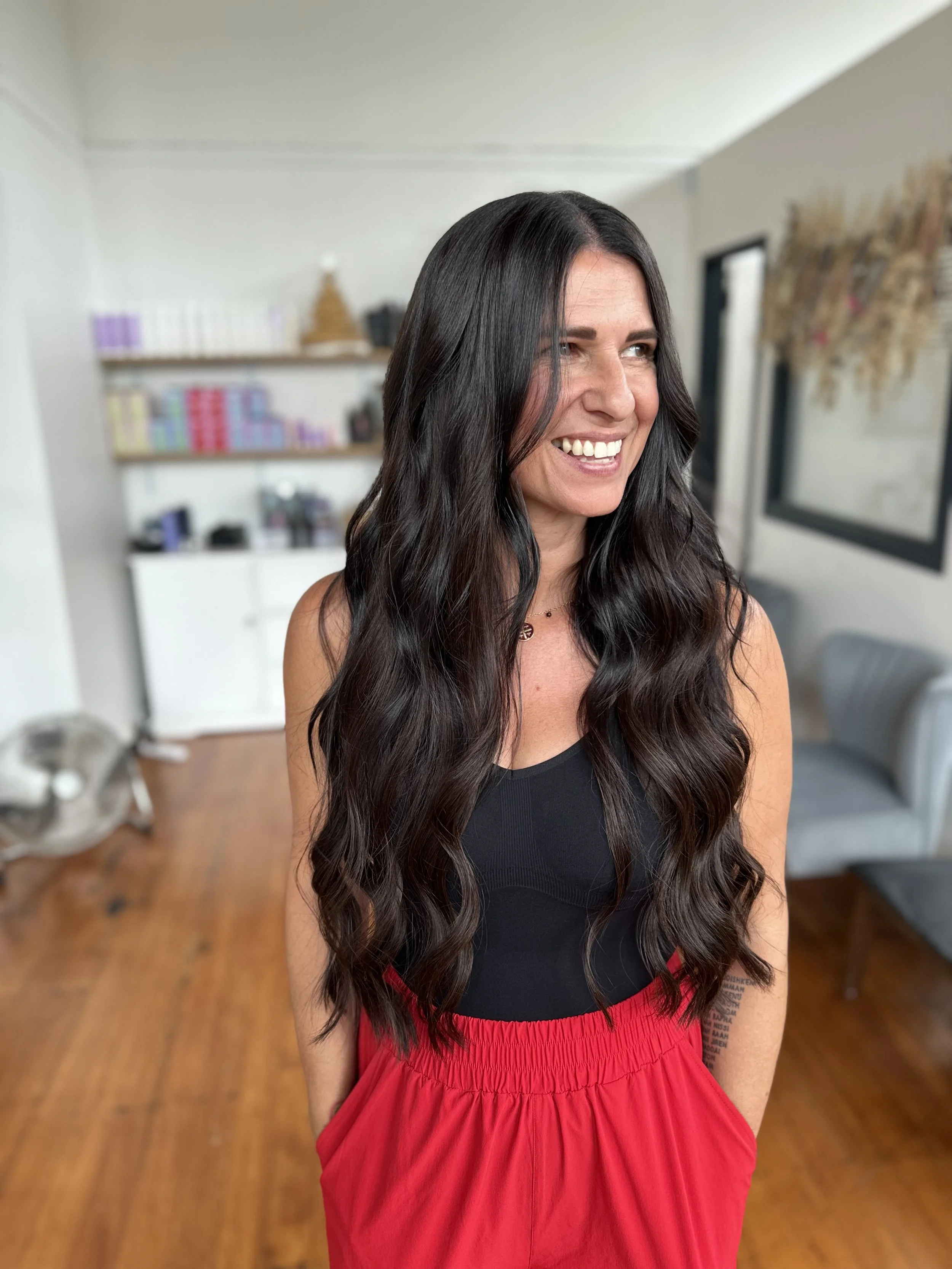 A woman with long, wavy black hair and a bright smile. She is wearing a black top and a red skirt, standing in a room with shelves filled with books and decorative items in the background.
