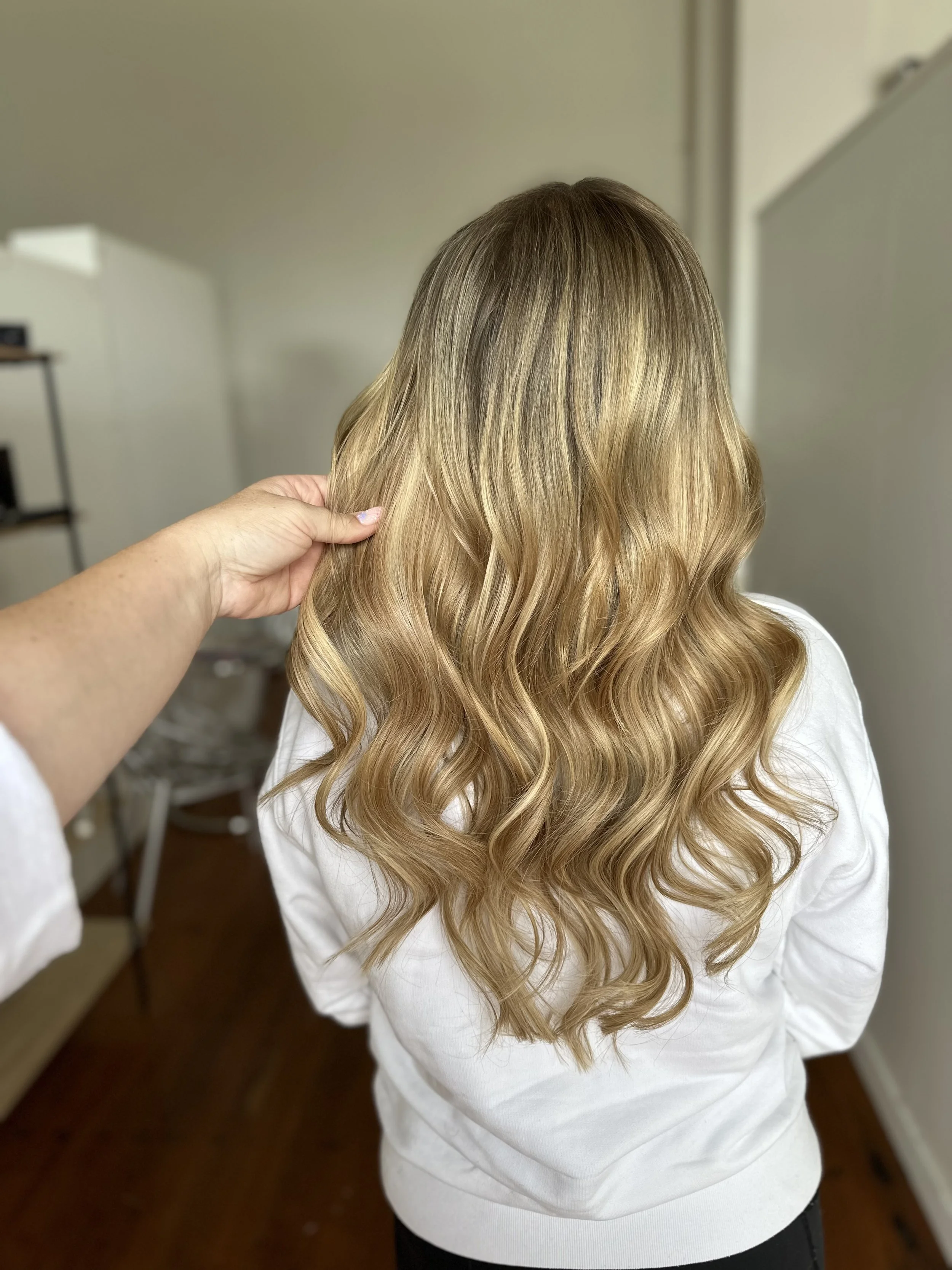 Woman with long, wavy blonde hair wearing a white shirt, seen from behind in a room.
