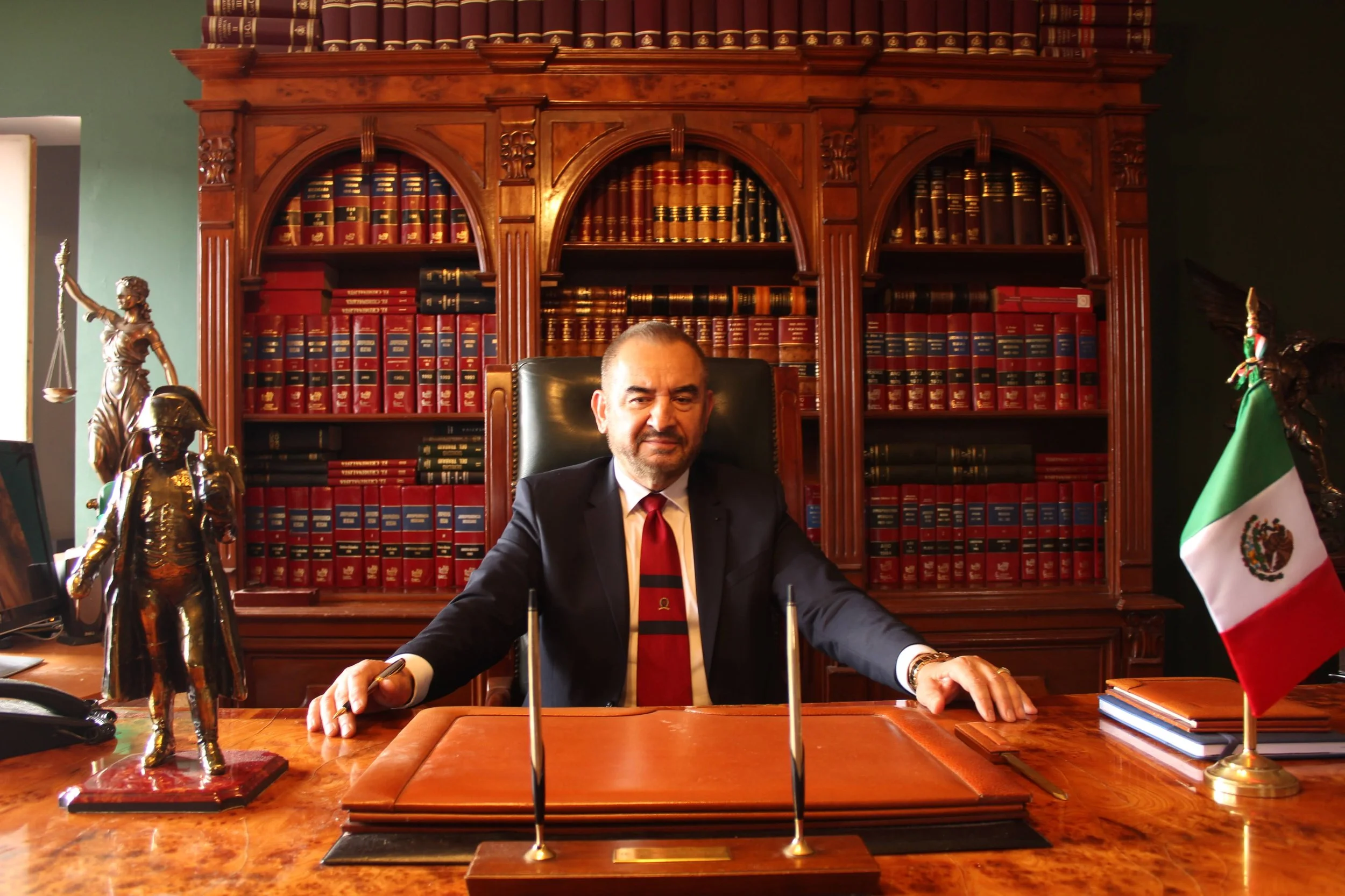 Un hombre sentado en una oficina con madera oscura, con estanterías llenas de libros y decoraciones de estilo clásico, además de banderas y estatuas, vestido con traje y corbata, en un entorno formal y elegante.