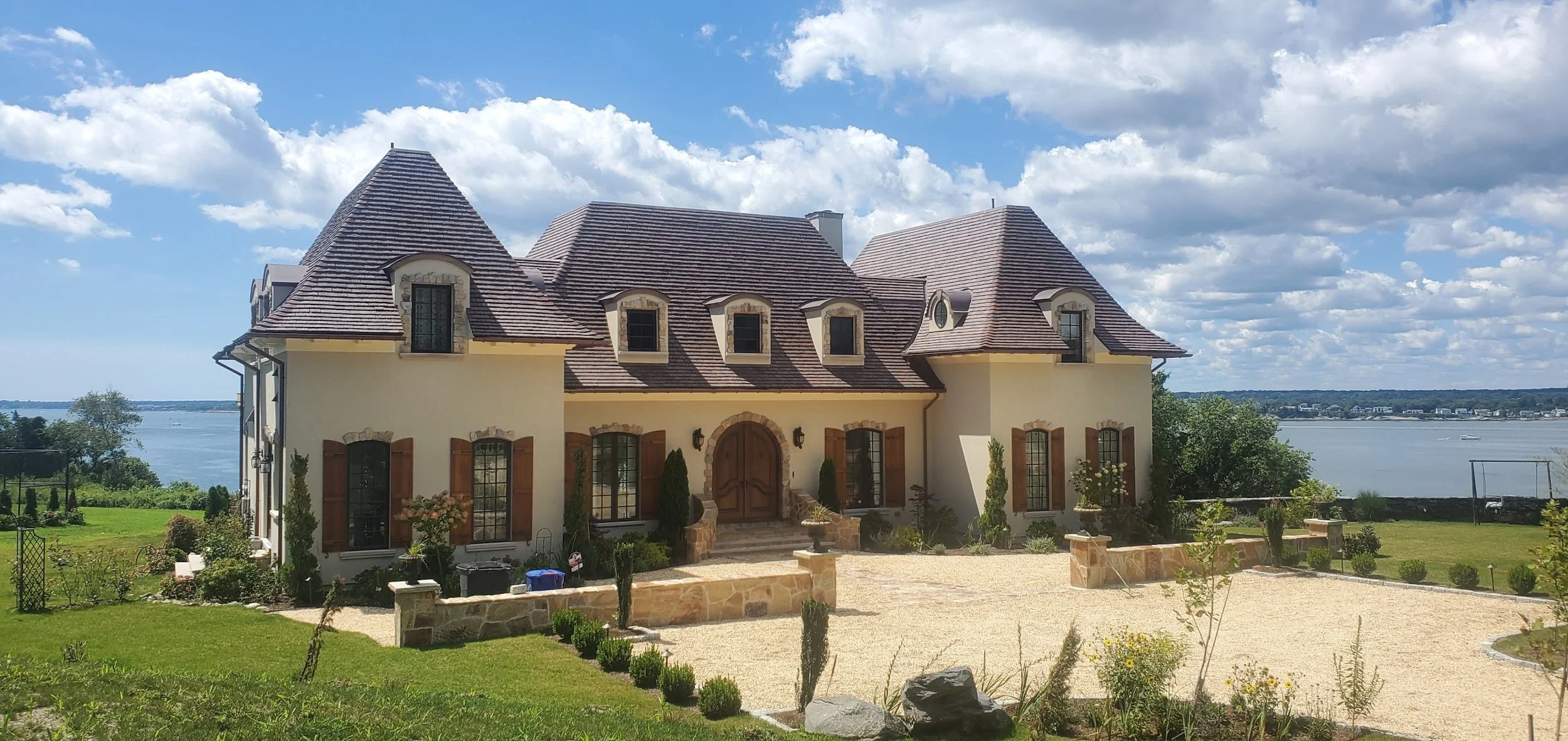 A large French-style mansion with a turreted roof, stone accents, and brown shutters, overlooking a lake under a partly cloudy blue sky.