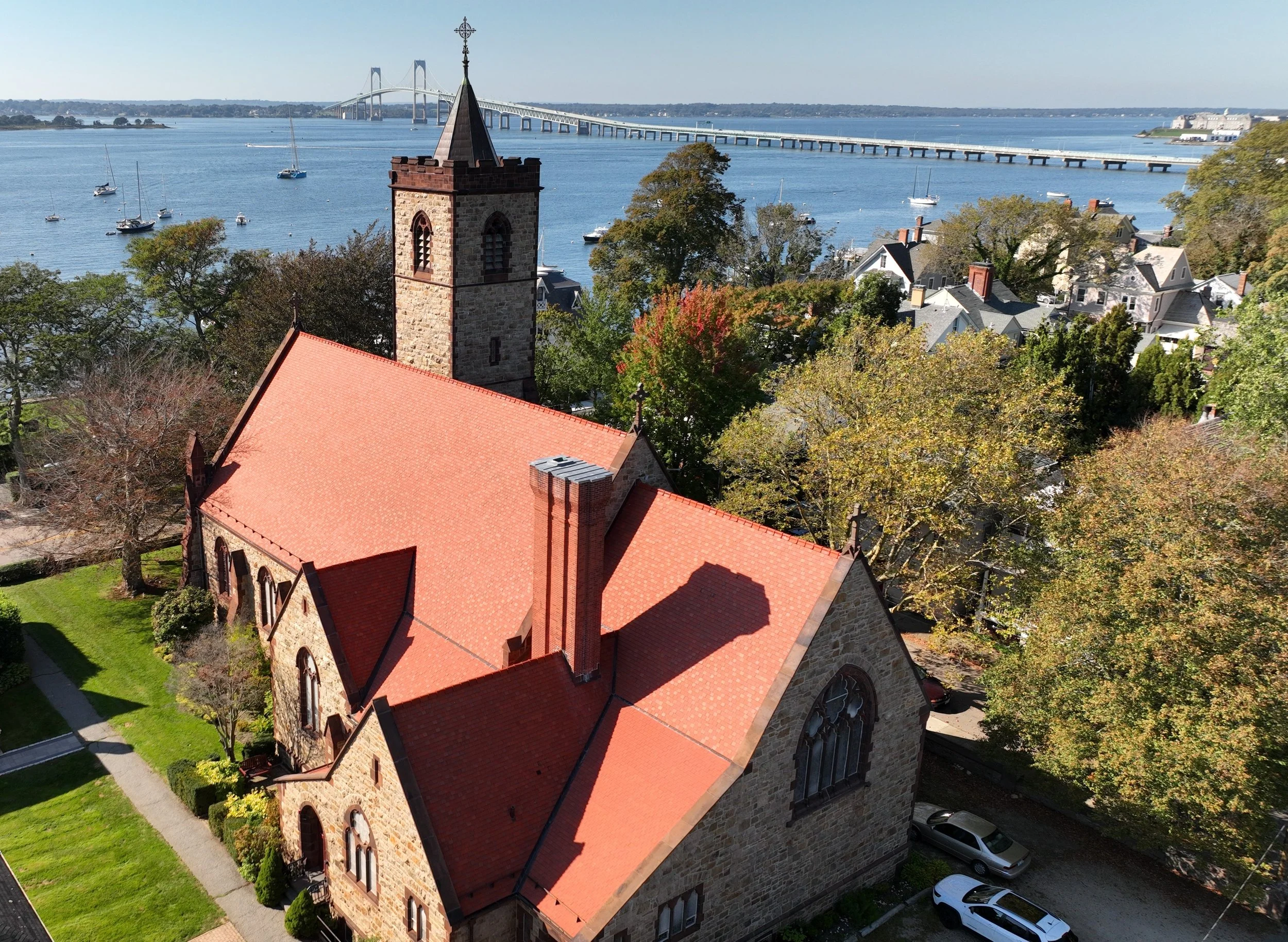 An aerial view of a stone church with a red-tiled roof, surrounded by trees and cars. The church has a tall steeple with a cross on top and overlooks a body of water with sailboats. In the background, there is a bridge spanning across the water.