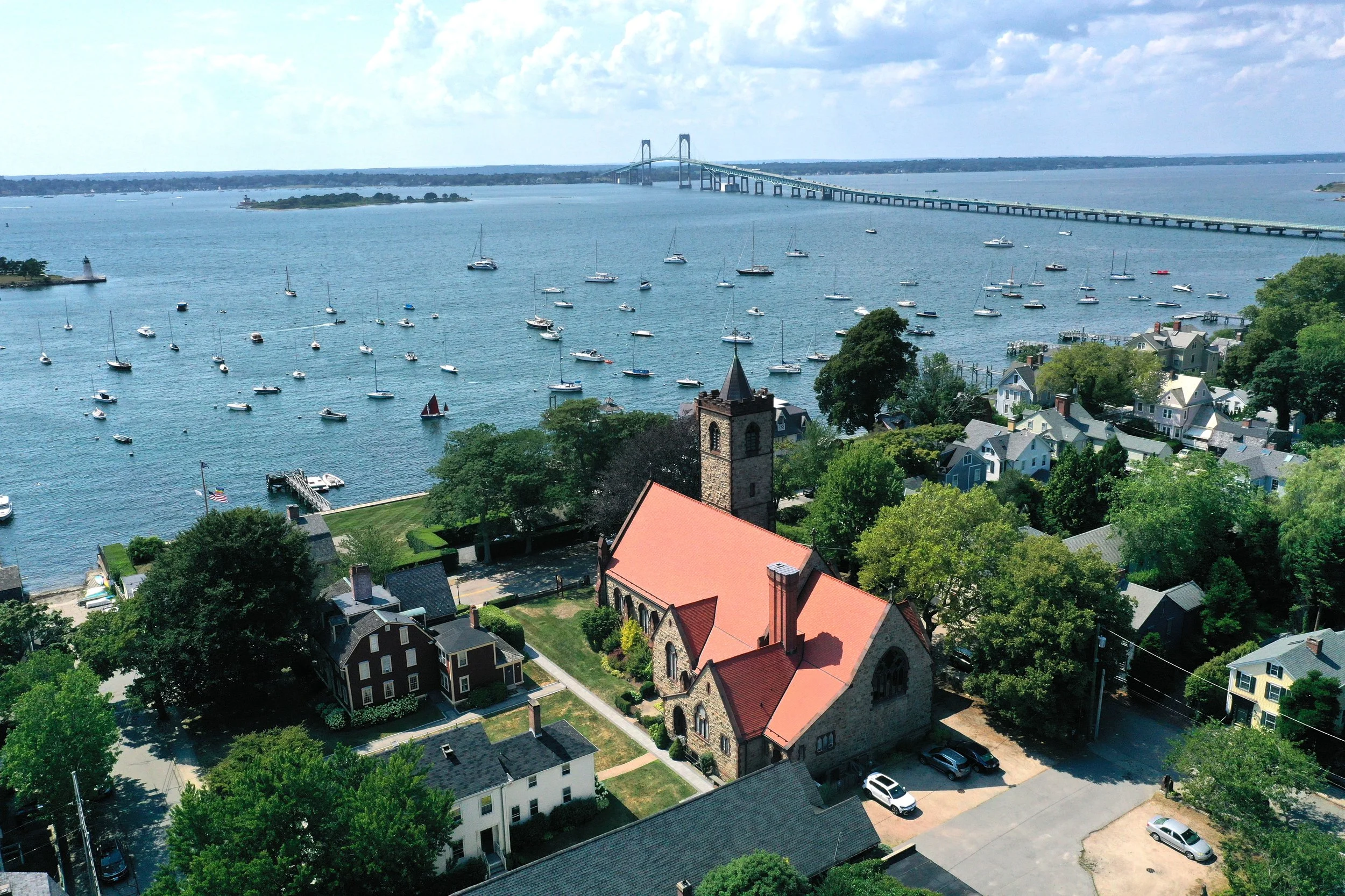 A scenic view of a harbor with numerous sailboats in the water, a bridge in the background, and residential buildings with green trees in the foreground.