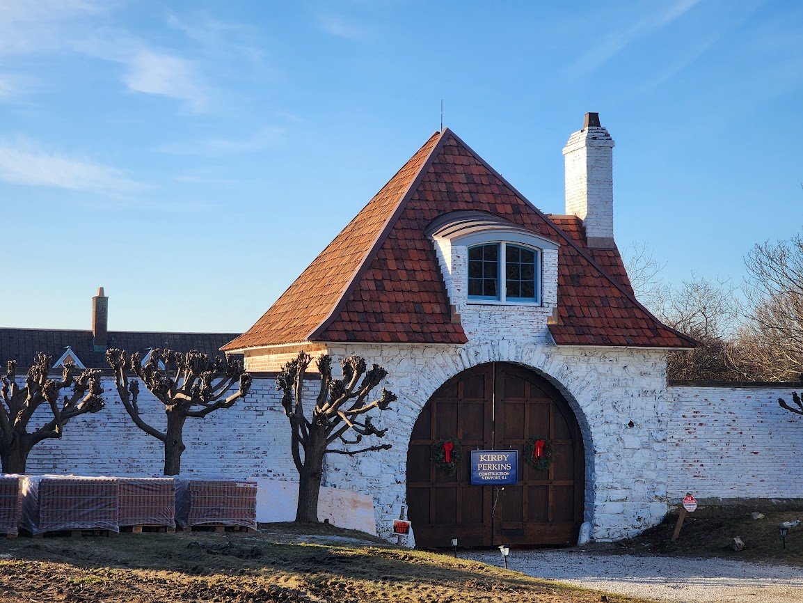 White brick wall with a large wooden gate decorated with Christmas wreaths, two pruned trees in front, and a house behind with a steeply pitched roof, chimney, and a dormer window.