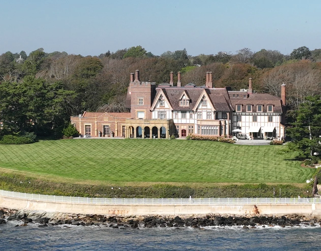 A large, historic mansion with a combination of brick and Tudor-style architecture sits on a grassy hill near the water.