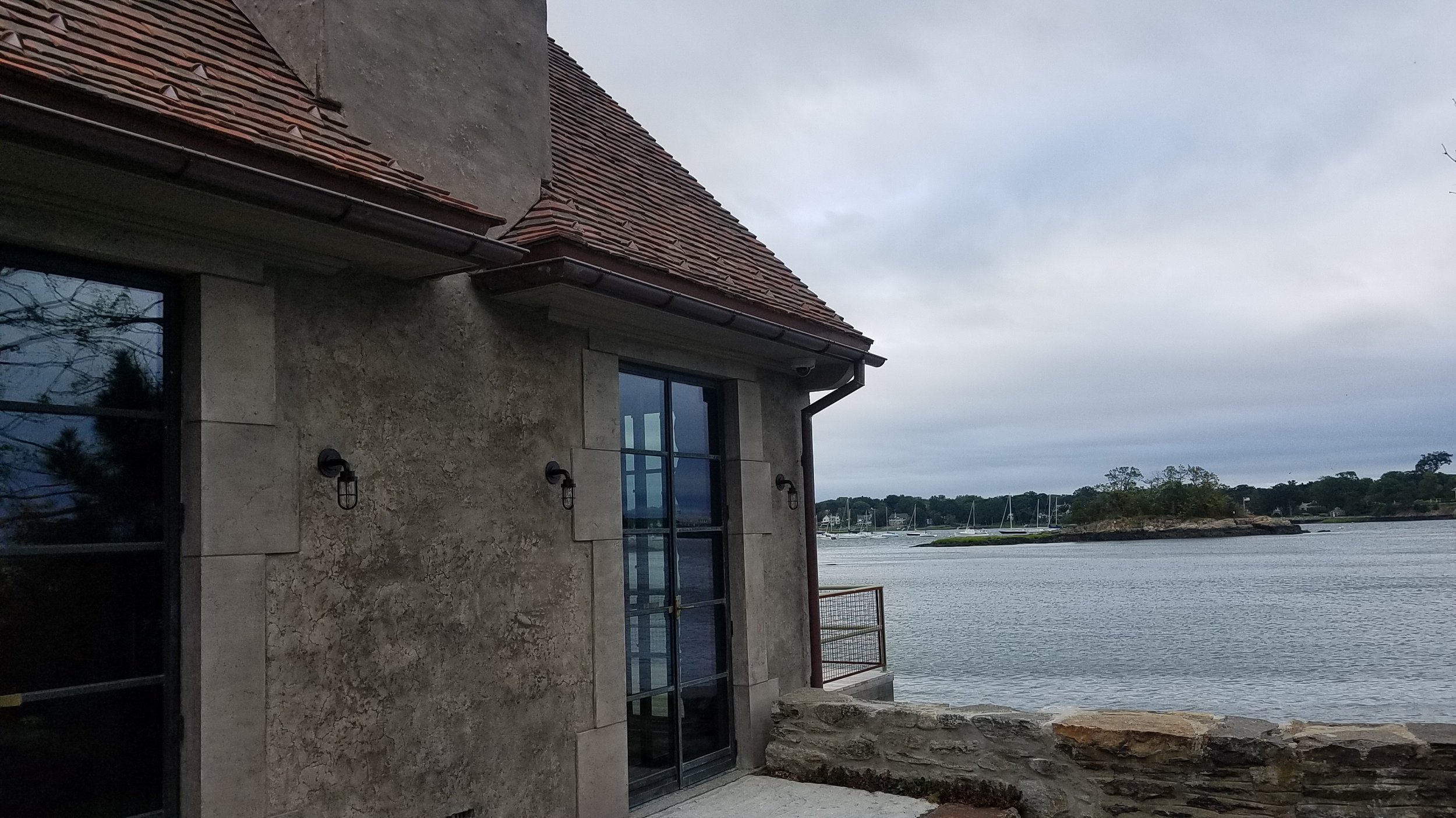 A house with stone walls and large windows near a body of water, with sailboats in the distance under a cloudy sky.