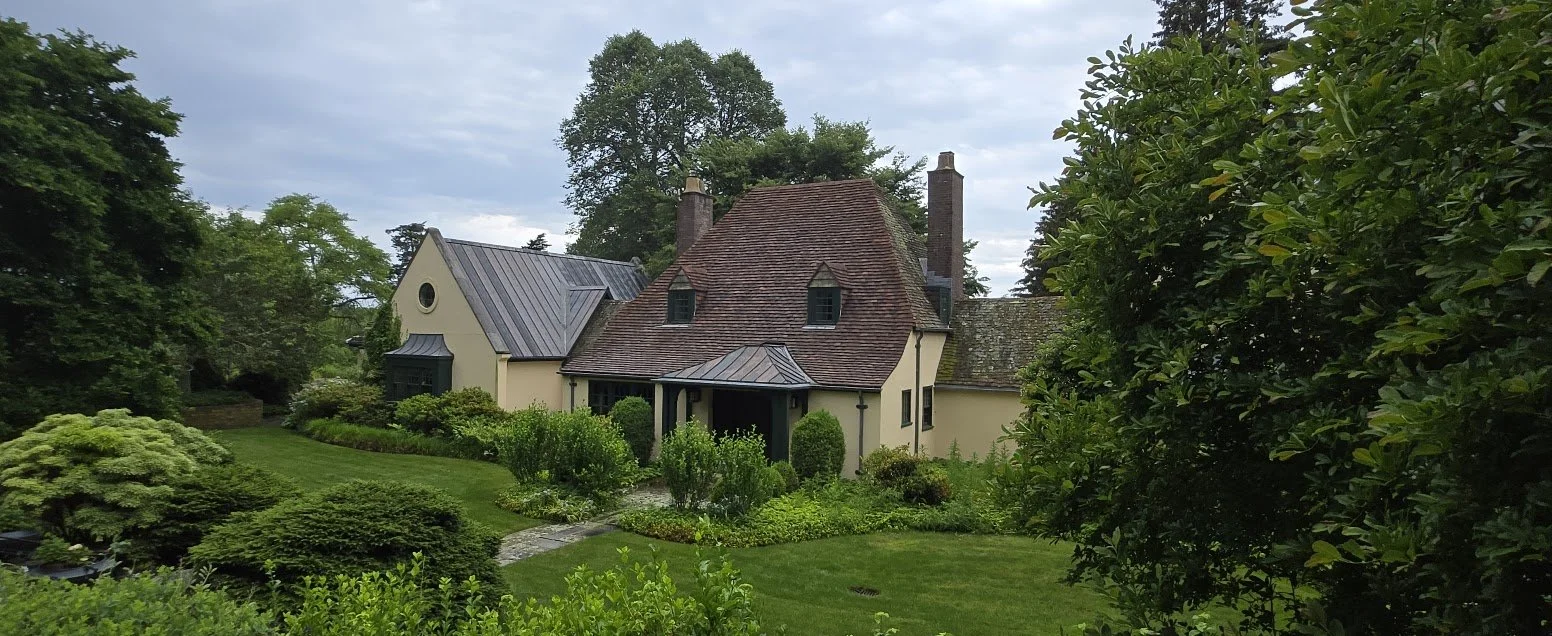 A house with a combination of different roof styles, surrounded by lush green trees, bushes, and a well-maintained lawn.
