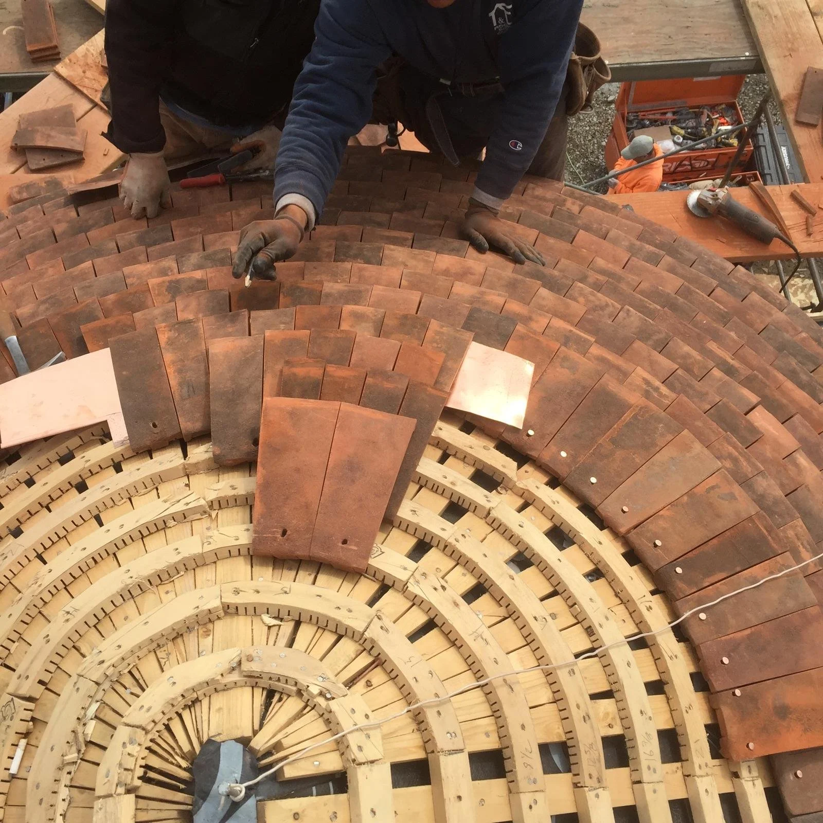 Workers assembling a curved wooden structure with reddish clay tiles on top, possibly a construction or architectural project.