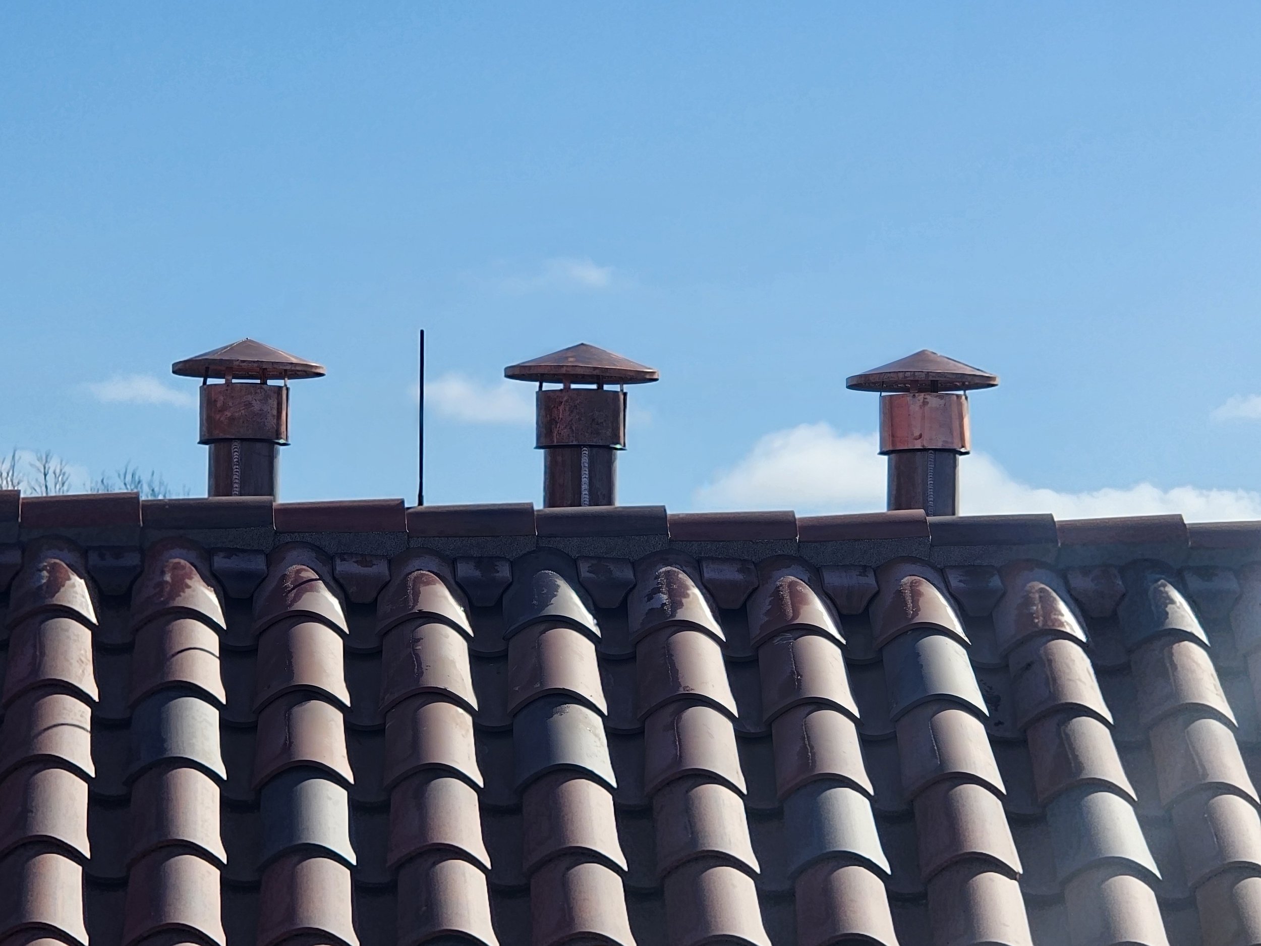 A rooftop with several chimney vents and a wire, with a background of blue sky and a few small clouds.