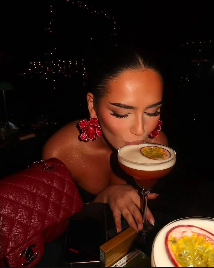 A woman with makeup and large red earrings is drinking a cocktail garnished with passion fruit, in a dimly lit setting.
