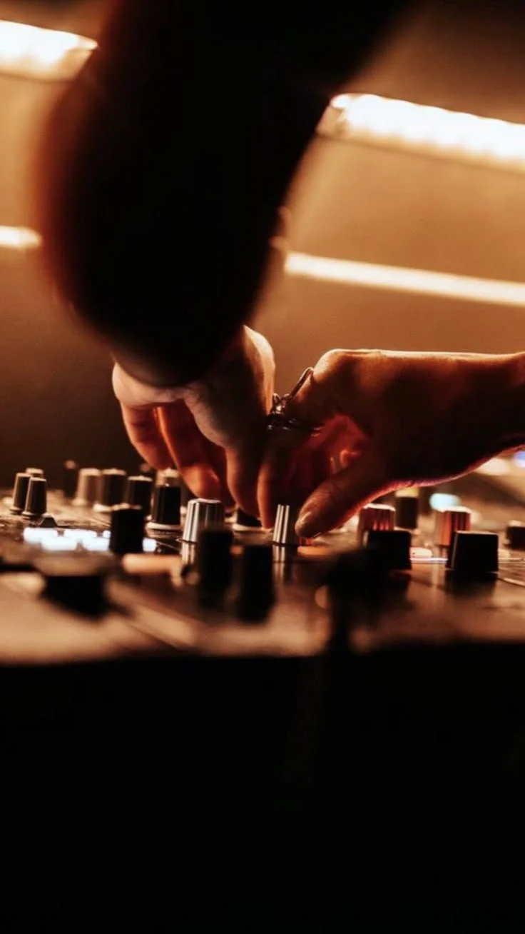 Close-up of hands adjusting knobs on a DJ mixing console in a dimly lit setting.