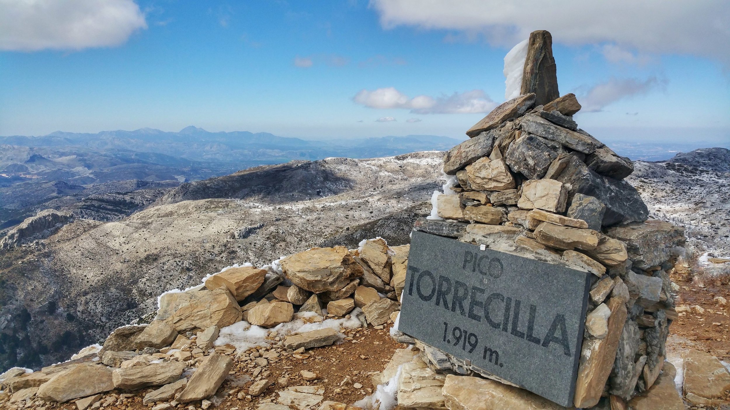 Formación de piedras en la cima de una montaña con vista panorámica de cadenas montañosas y cielo con nubes, señal que indica la cumbre con una altura de 1919 metros.
