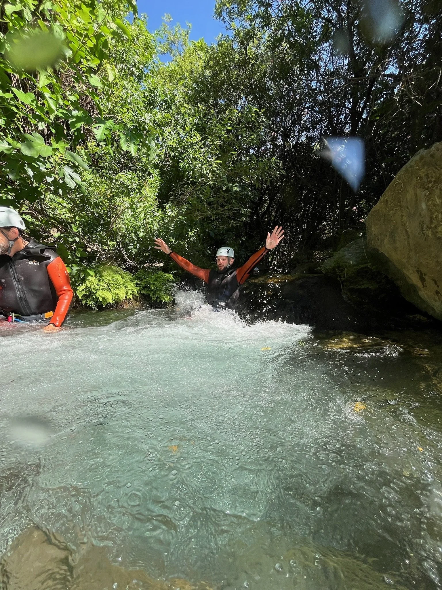 Dos personas con cascos de protección y trajes de neopreno disfrutan de un descenso en kayak por un río con mucha agua y rodeado de vegetación.