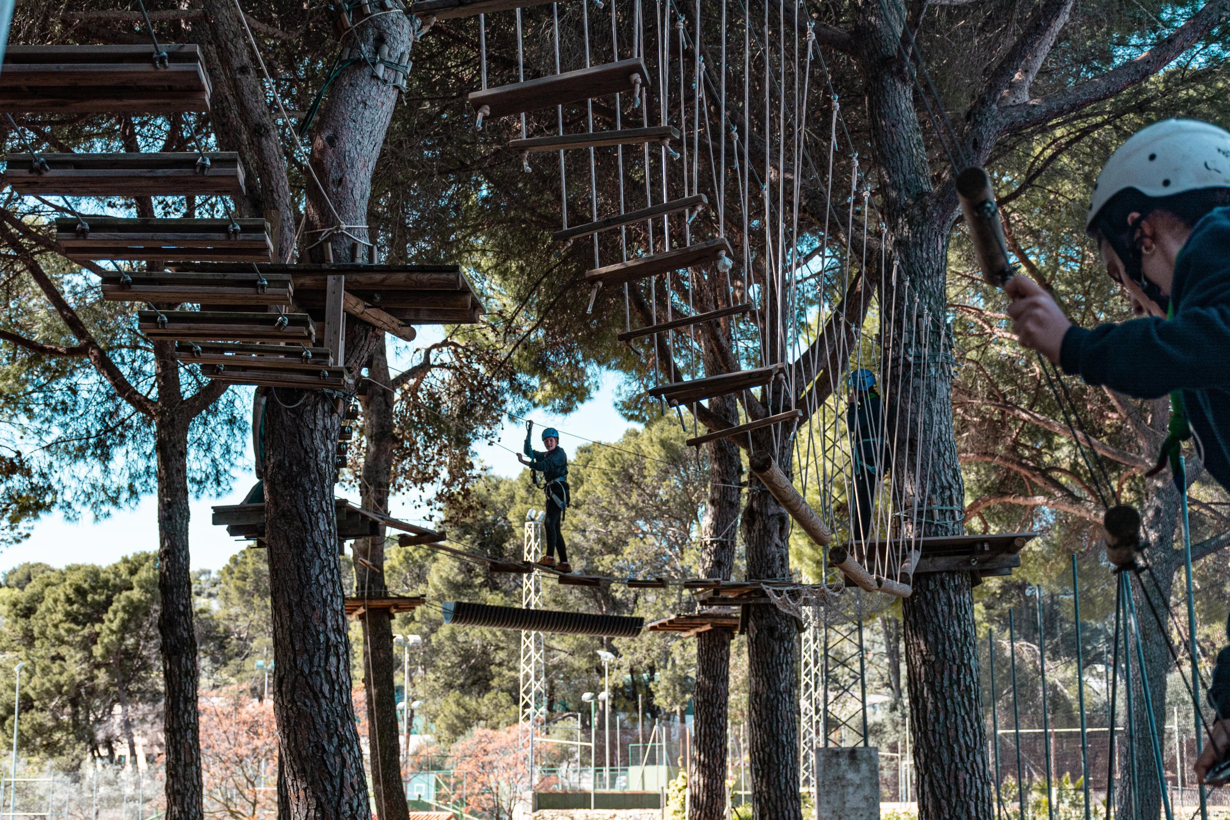 Personas en un parque de aventuras en los árboles, algunos caminando en puentes colgantes y otros trabajando en la cuerda con casco.