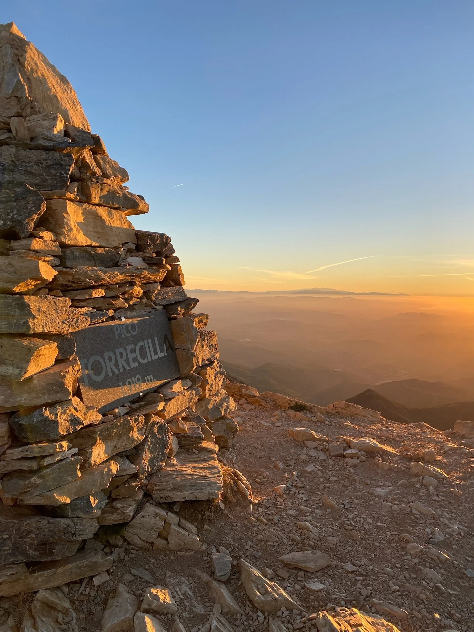 Vista de una montaña rocosa al atardecer con un cielo despejado y un cartel que indica la cima, en tonos dorados por el sol poniente.