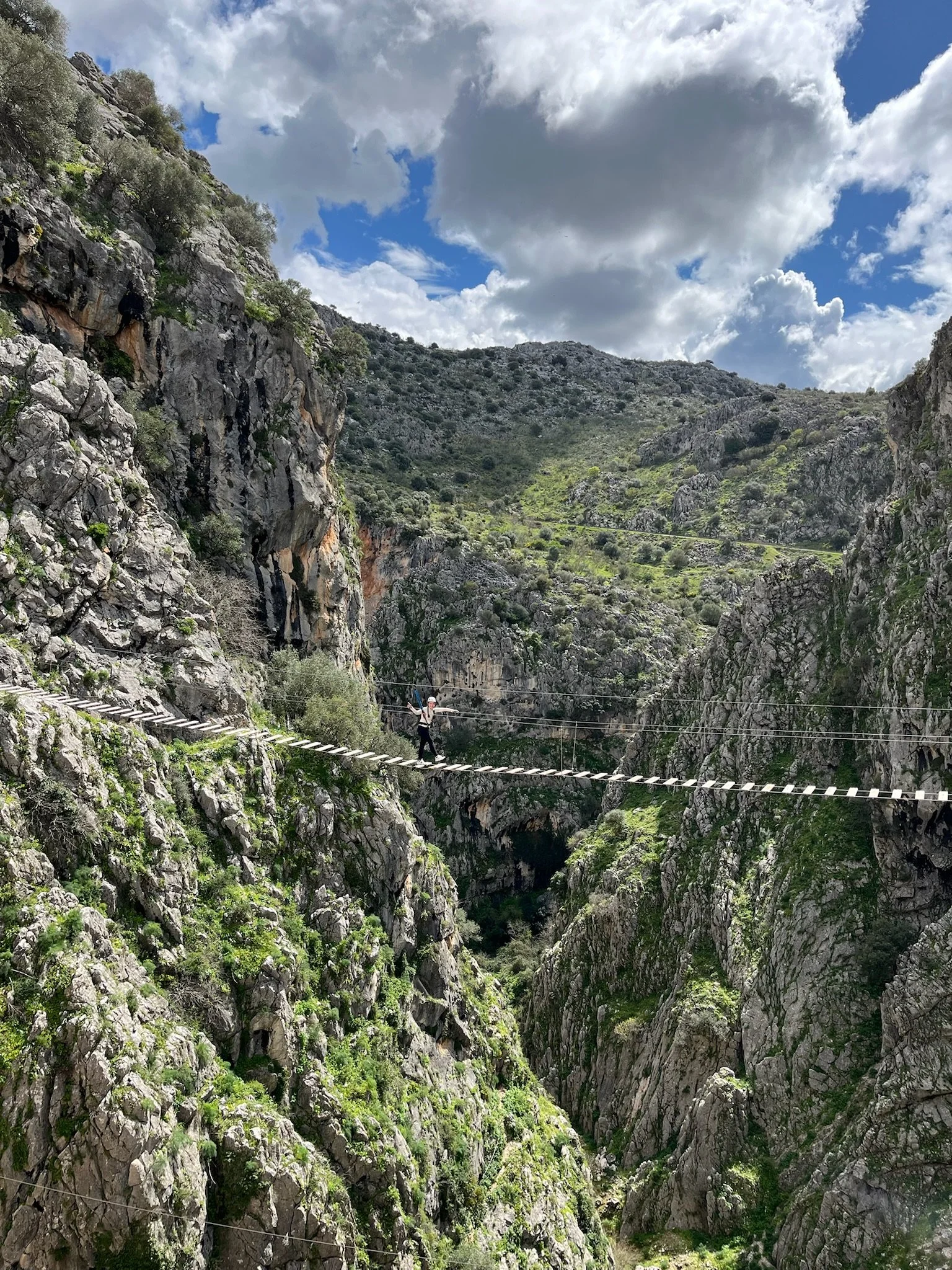 Persona cruzando un puente colgante en un cañón rodeado de montañas rocosas y vegetación, cielo con nubes grandes y cielo parcialmente azul.