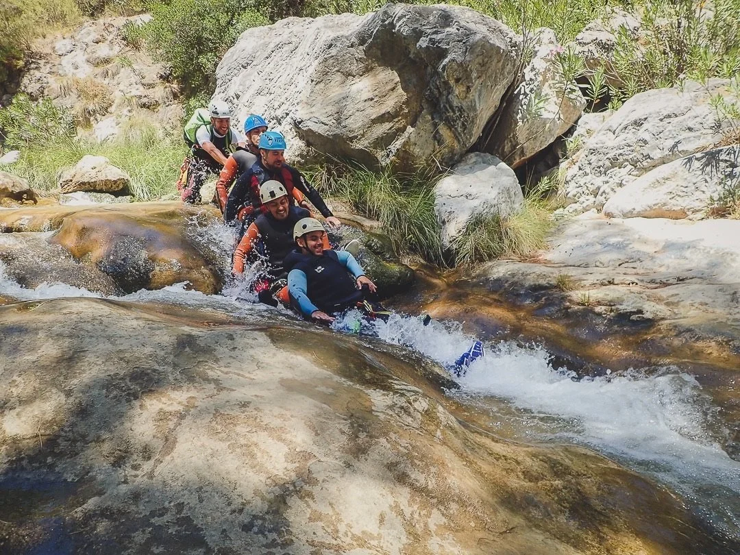 Grupo de seis personas haciendo rafting en un río con rocas grandes y vegetación.