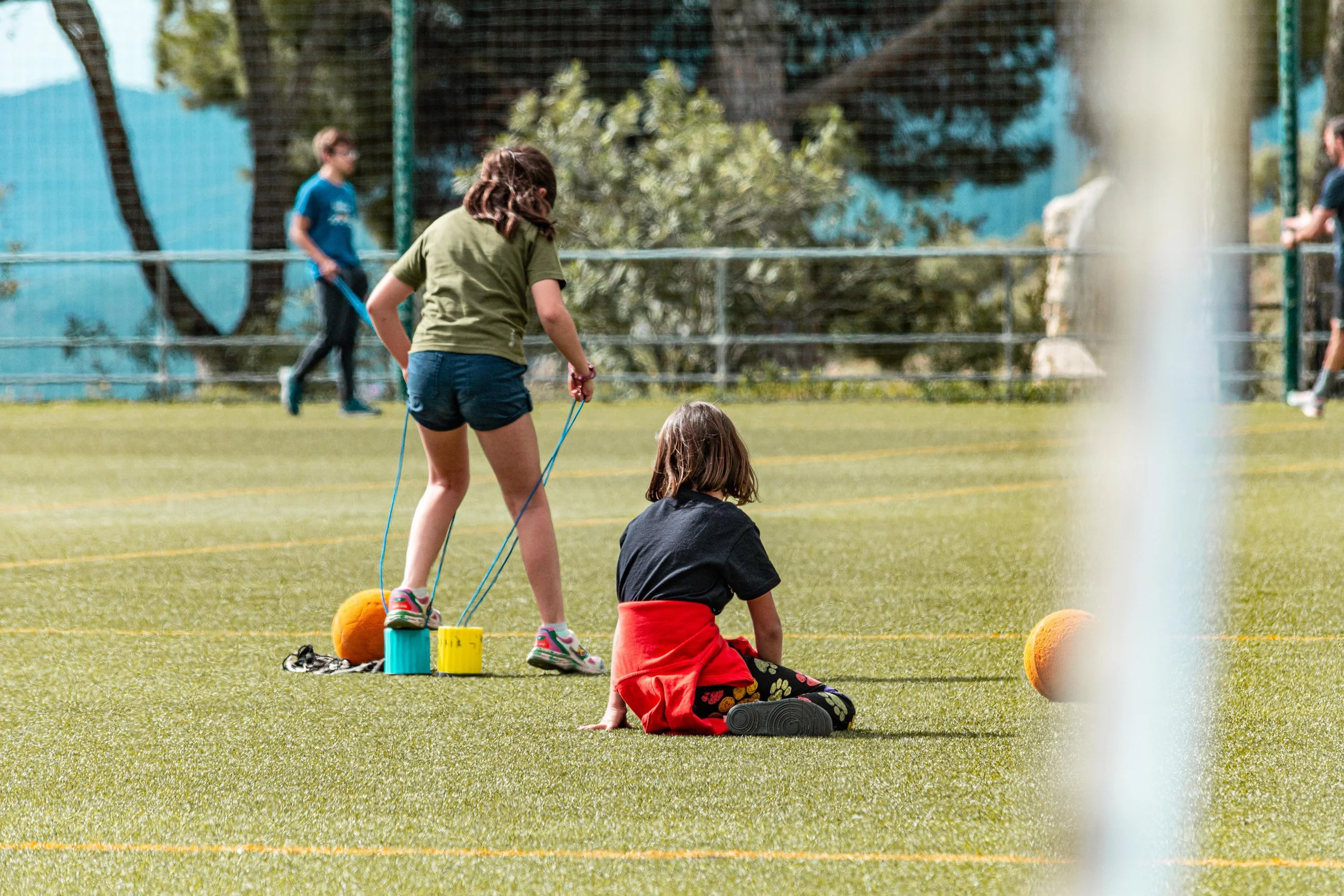 Niños y niñas jugando en un campo de futsal, algunos sentados y otros de pie, rodeados de un área con césped artificial, en un ambiente al aire libre con árboles y malla perimetral.