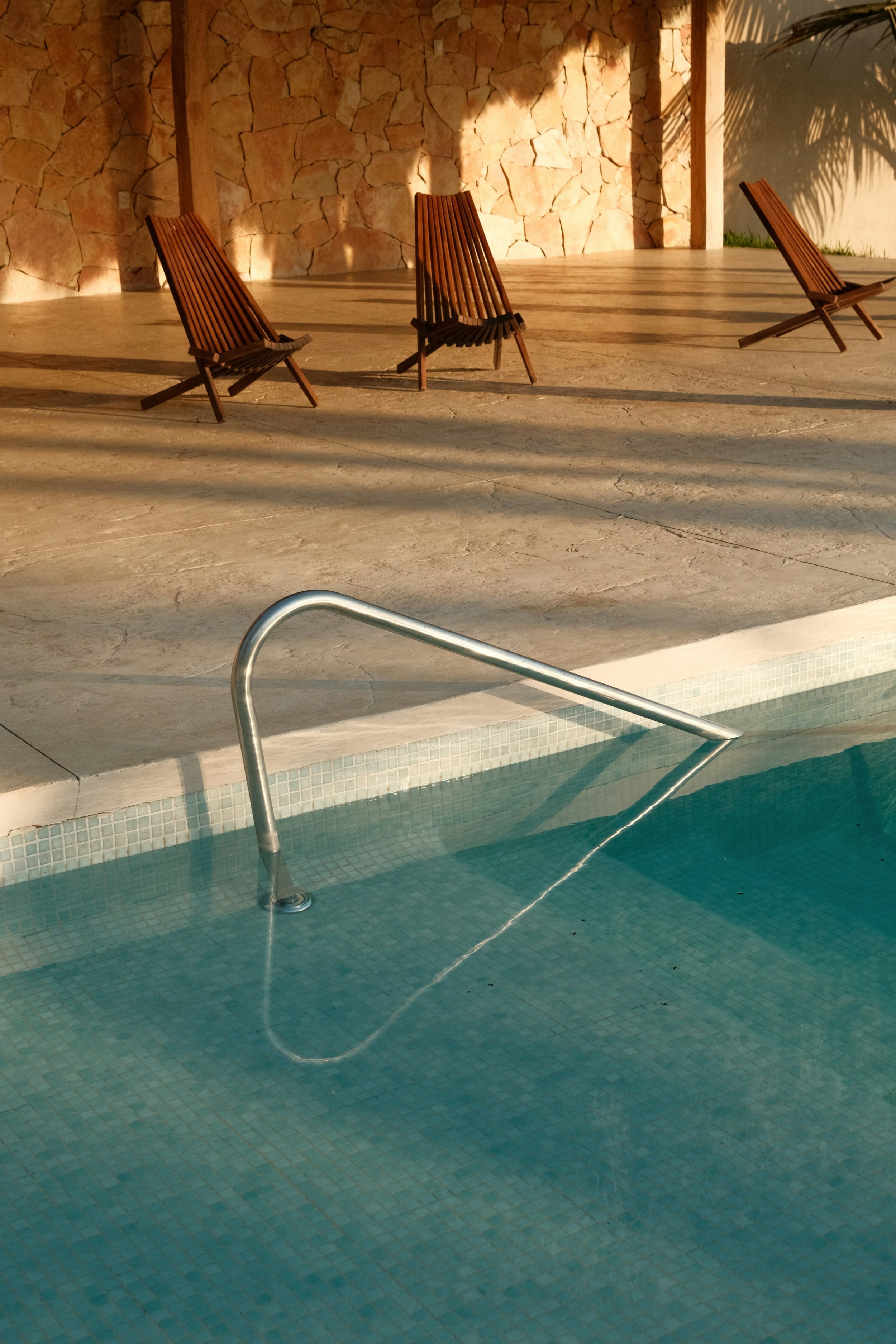Three wooden lounge chairs on a beige stone patio near a swimming pool, with a stone wall and shadow of a palm tree in the background.