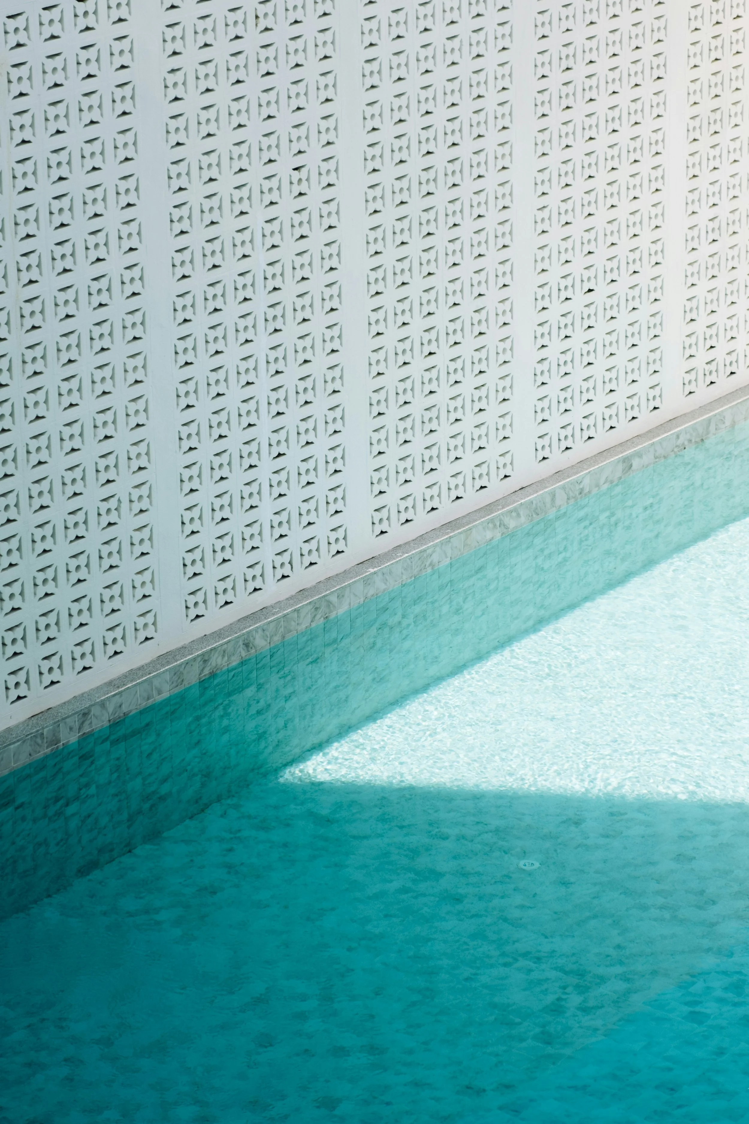 A white lattice wall next to a swimming pool filled with blue water, with sunlight casting shadows on the pool's surface.