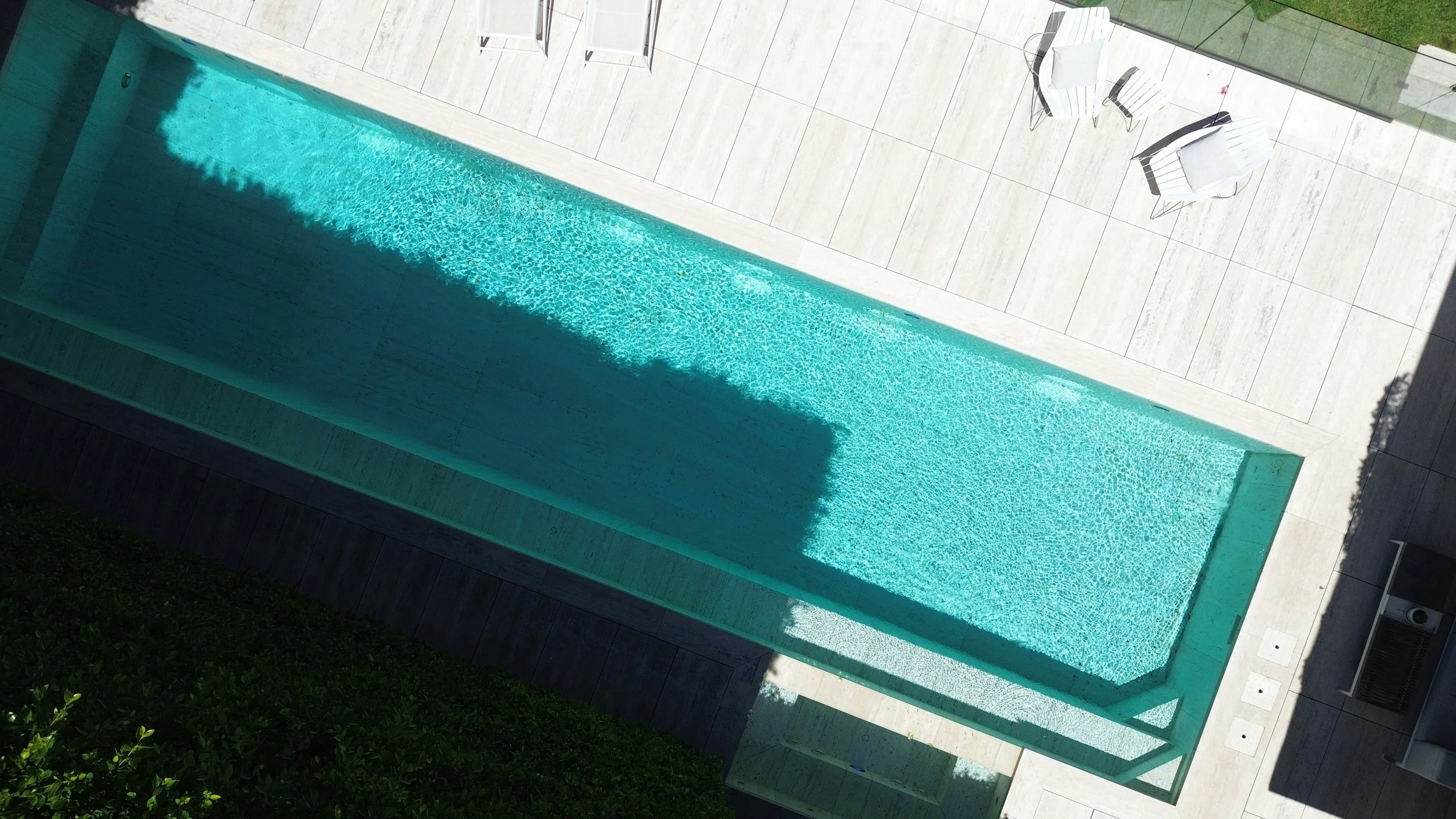 A modern outdoor swimming pool with turquoise water, surrounded by a light-colored tiled deck with white chairs and a table, and a glass fence with greenery outside.