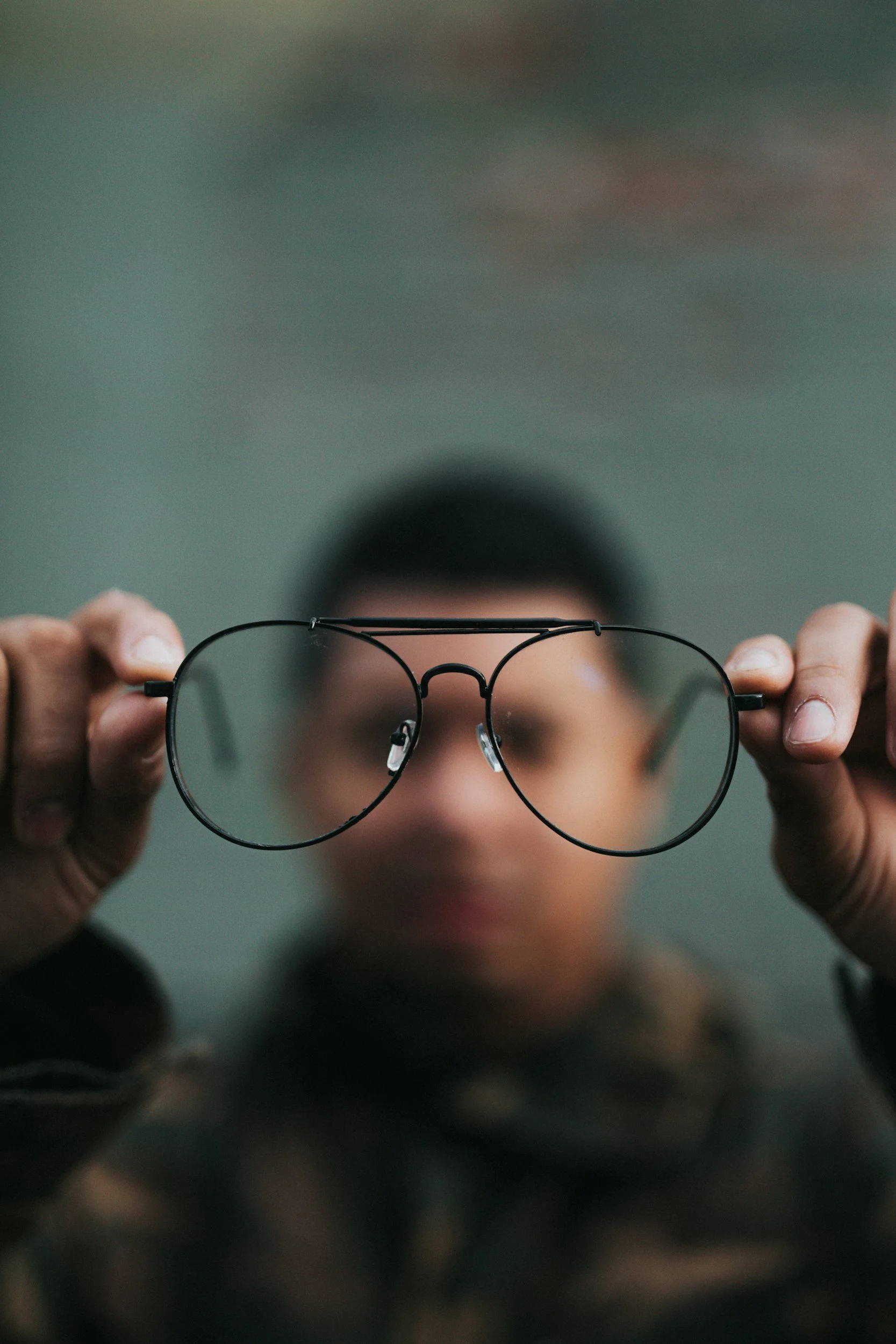 Person holding sunglasses close to the camera, with focus on the sunglasses and blurry face and background.
