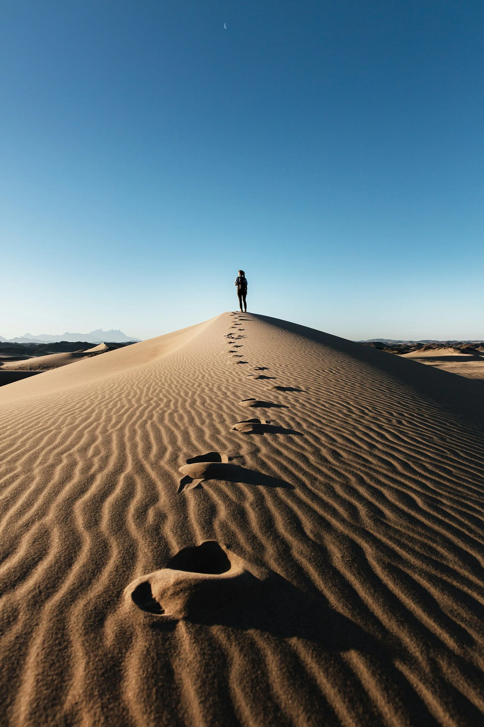 A person standing on a sand dune in a desert, with footprints leading up to them, under a clear blue sky with a crescent moon visible.