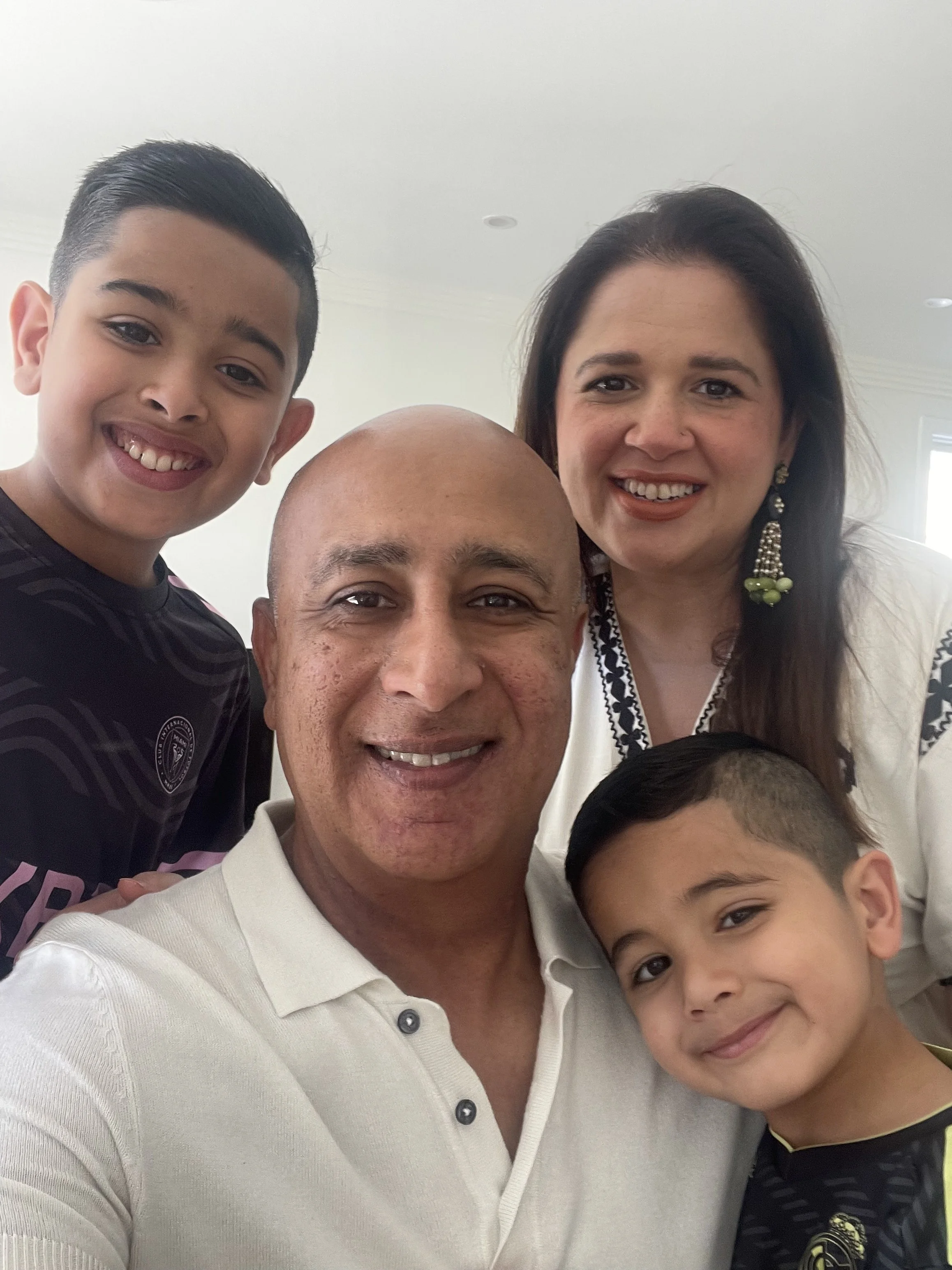 Group photo of four people, including a smiling young boy with dark hair and an adult man with a shaved head, taken indoors with a white ceiling and windows in the background.