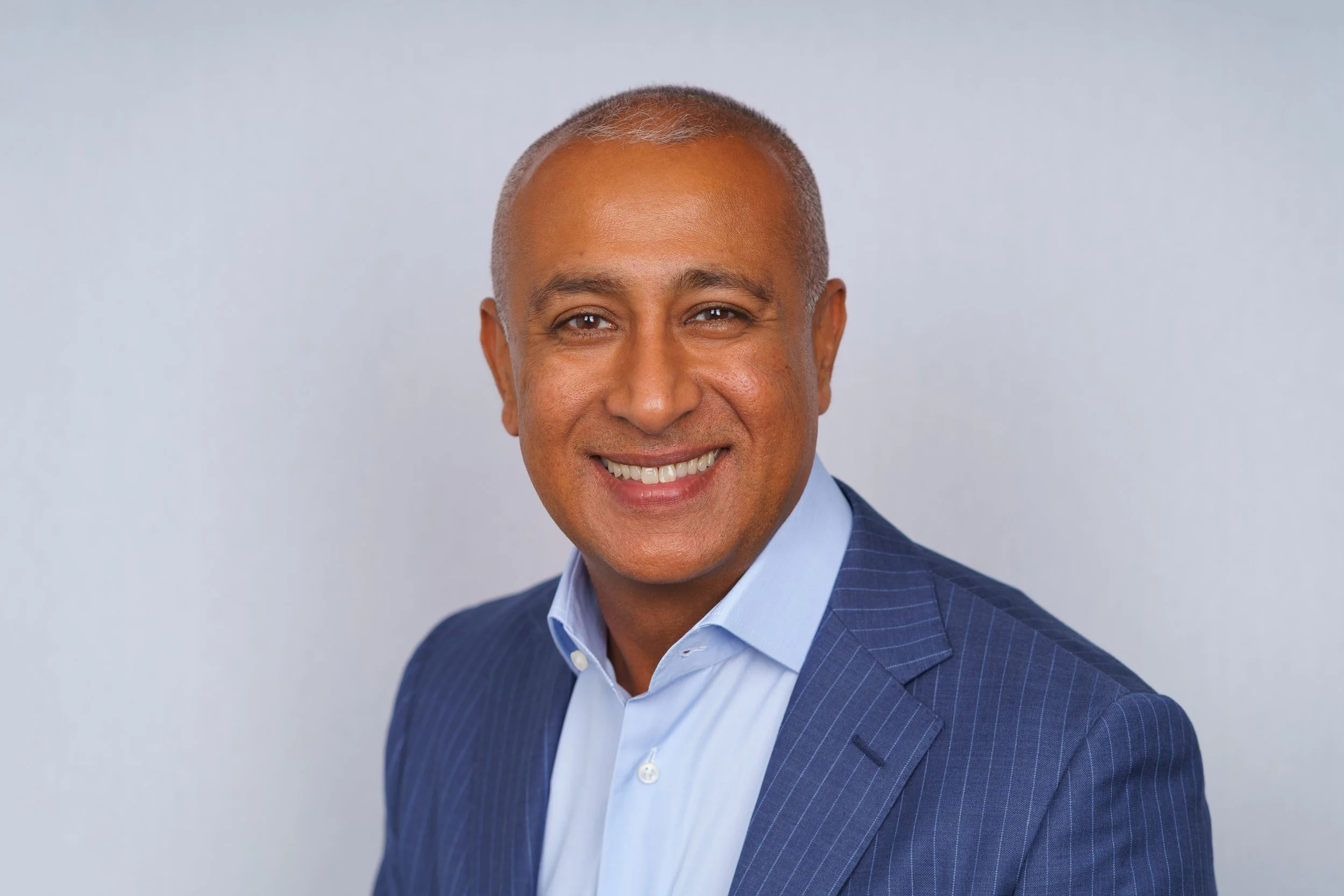 A middle-aged man with short gray hair smiling and wearing a blue suit and light blue shirt, posing against a plain light gray background.