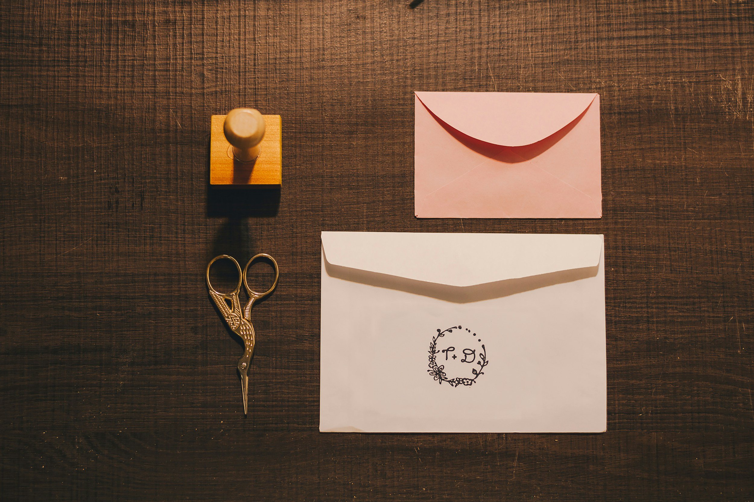 Flat lay of a wooden table with a rubber stamp, scissors, blank white paper, pink paper with a curved top and folded bottom, and a piece of paper with a floral wreath design.