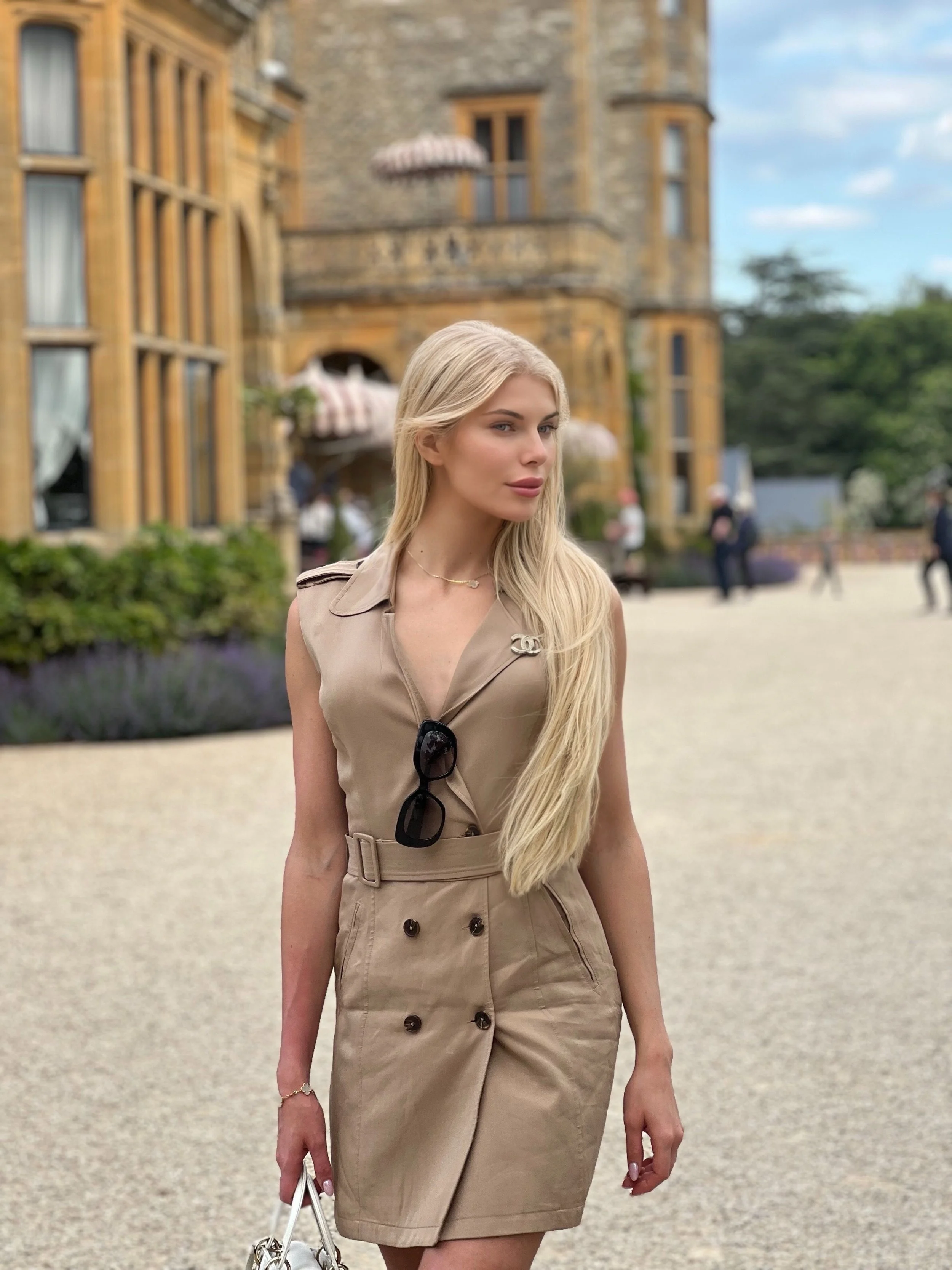 A young woman with long blonde hair wearing a beige sleeveless trench dress, sunglasses hanging from the front, and a bracelet, holding a white handbag, standing outdoors at Manor Estelle.
