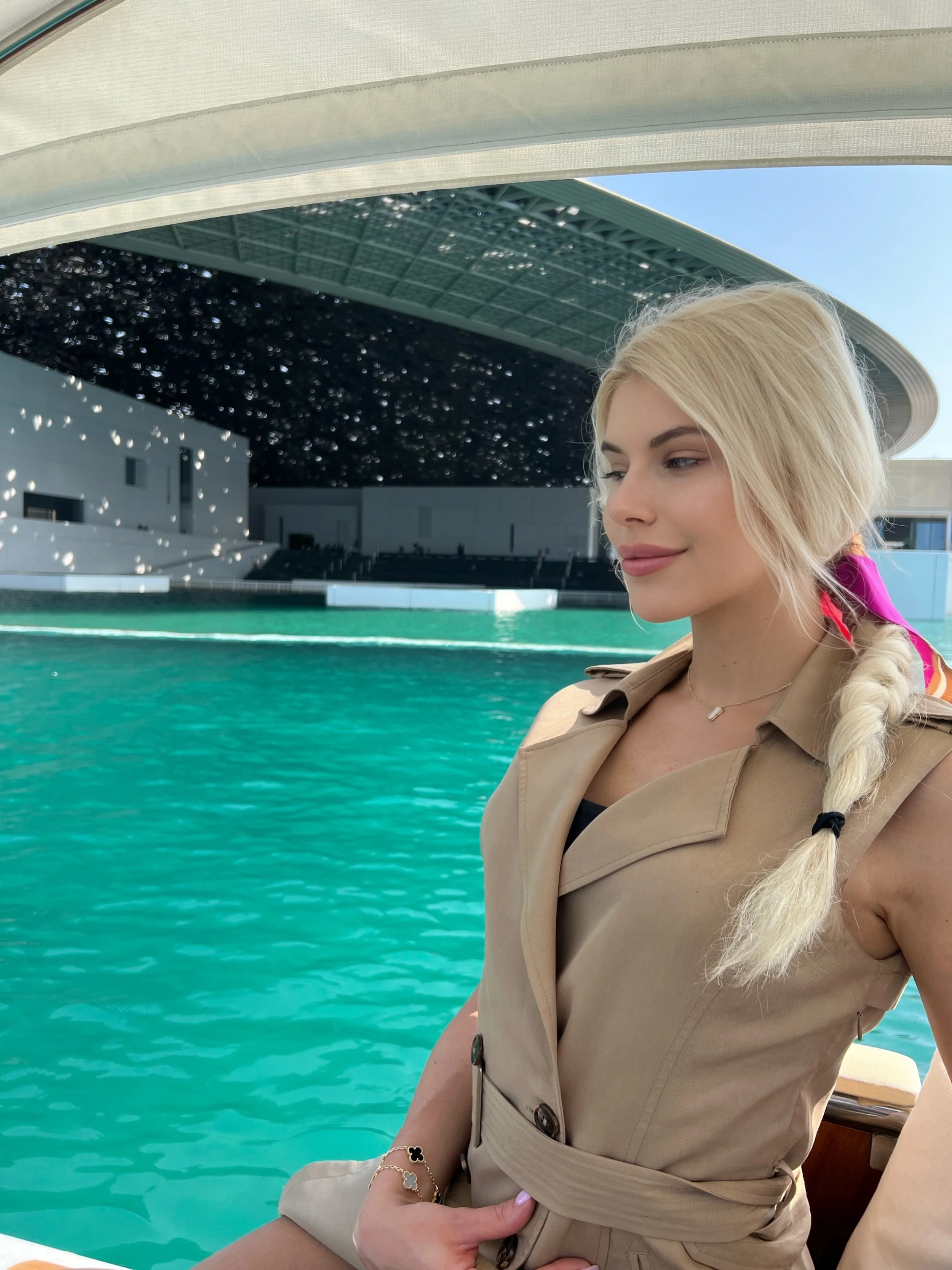 Young woman with blonde hair styled in a braid, wearing a beige trench coat and jewelry, sitting by a turquoise body of water in Louvre Abu Dhabi.