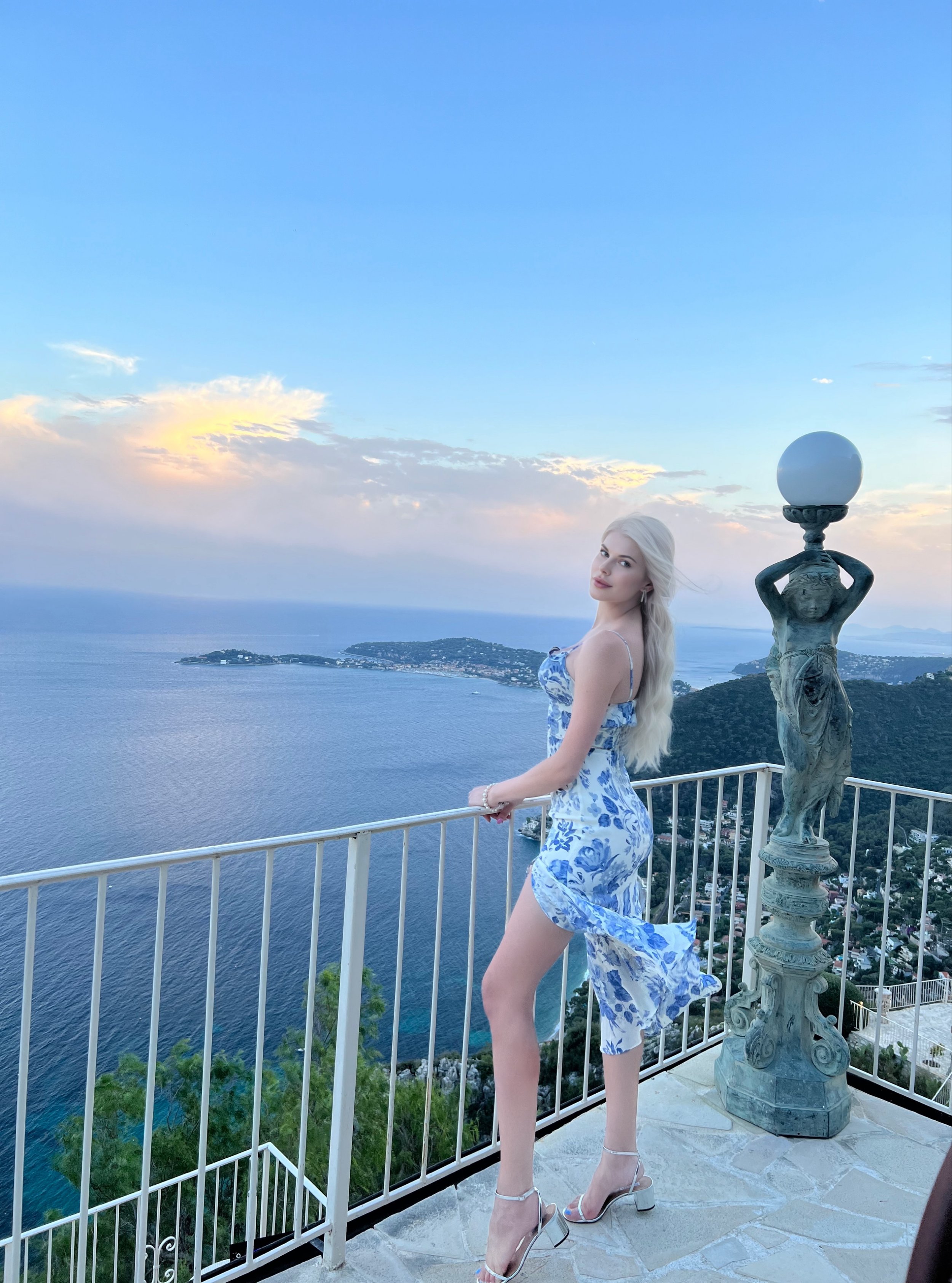A woman with long blonde hair in a blue and white floral dress standing on a balcony at Château de La Chèvre d'Or.