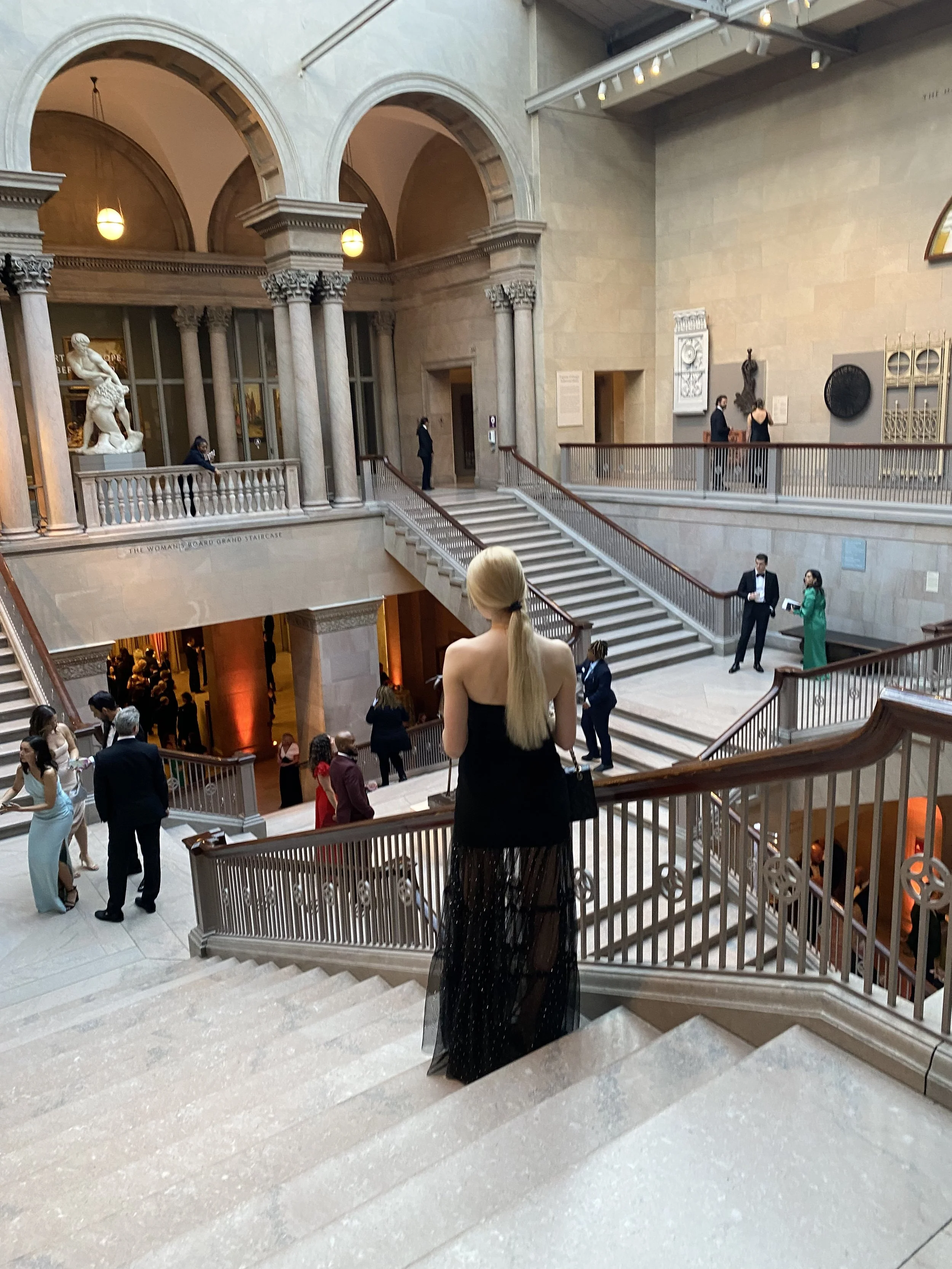 An elegant woman with long blonde hair and a black dress standing on a staircase of the Art Institute of Chicago, with people dressed formally around her, including men in tuxedos and women in evening gowns, during a social or formal event.