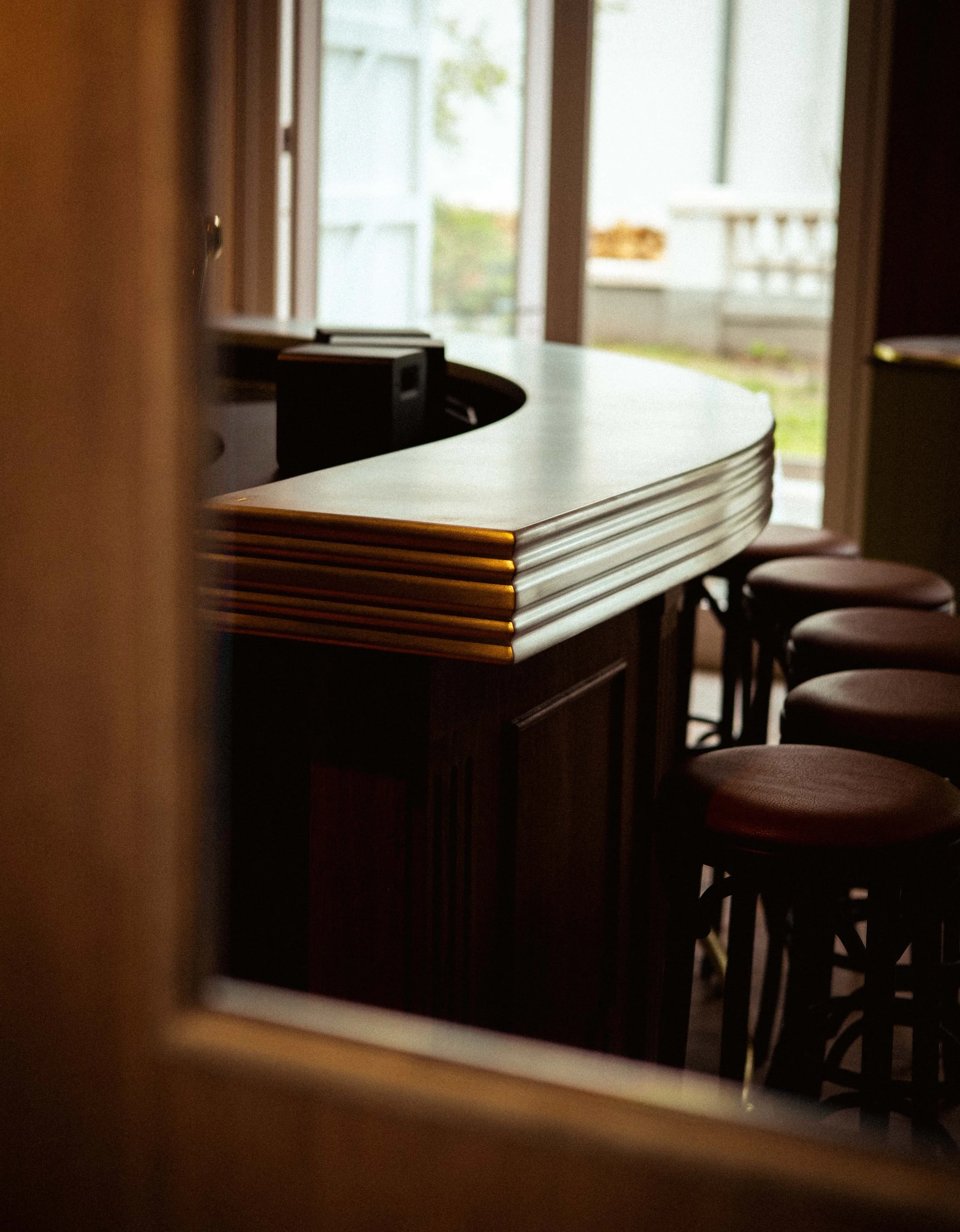 Binnenkant van een bistro, deels zichtbaar door een raam, met een hoek van een tafel en stoelen, en een deur die uitkomt op een balkon of veranda met een witte reling.