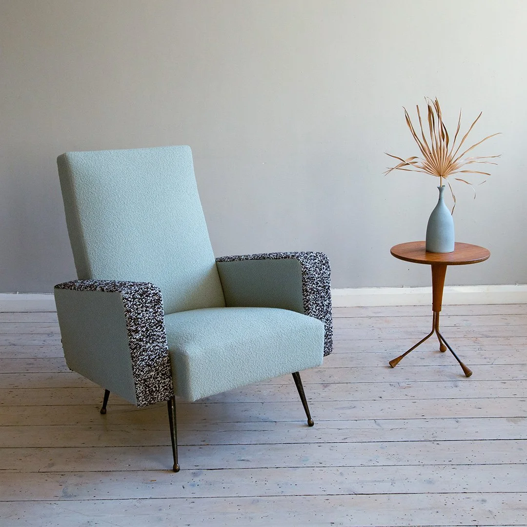 A light blue French mid-century modern armchair with black and white textured arms, placed on a white wooden floor next to a round wooden side table with a blue vase holding dried palm leaves against a plain neutral wall.