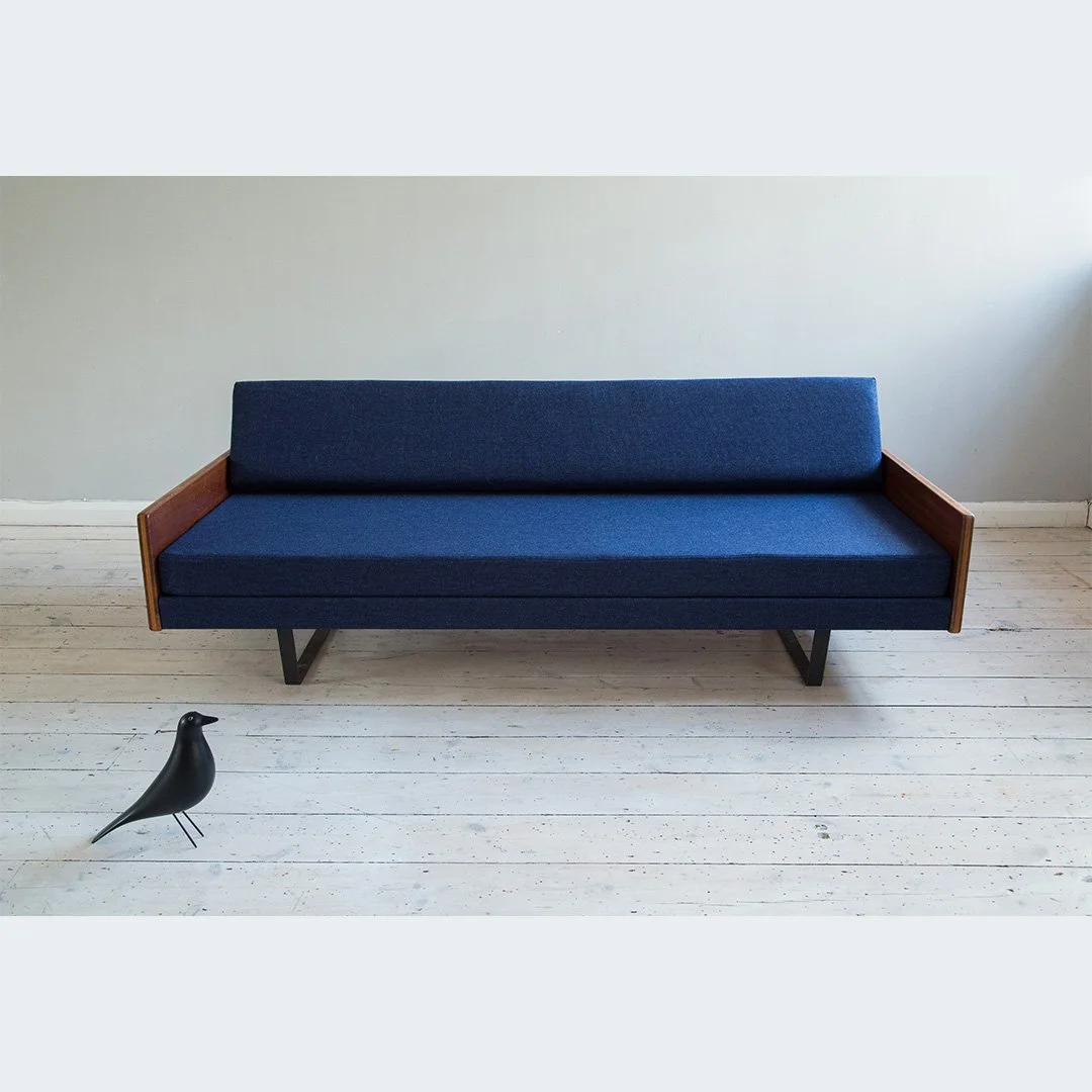 A modern blue Robin Day sofa with wooden armrests and black legs, positioned against a light-coloured wall on a white wooden floor, with a black decorative bird sculpture on the floor in front of it.