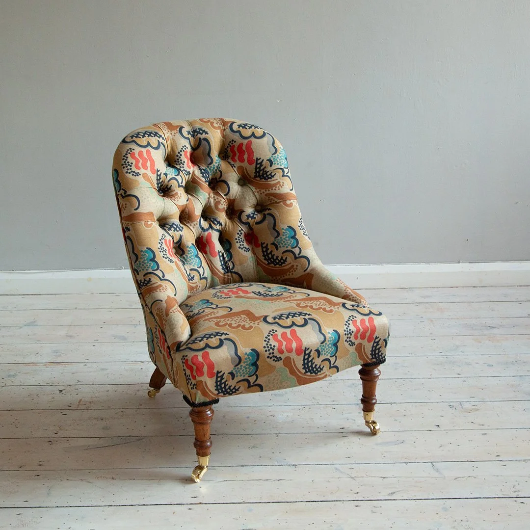A Victorian armchair with a colourful, patterned fabric by Duncan Grant from Charleston, wooden legs with brass castors, set against a plain grey wall and white wooden floor.