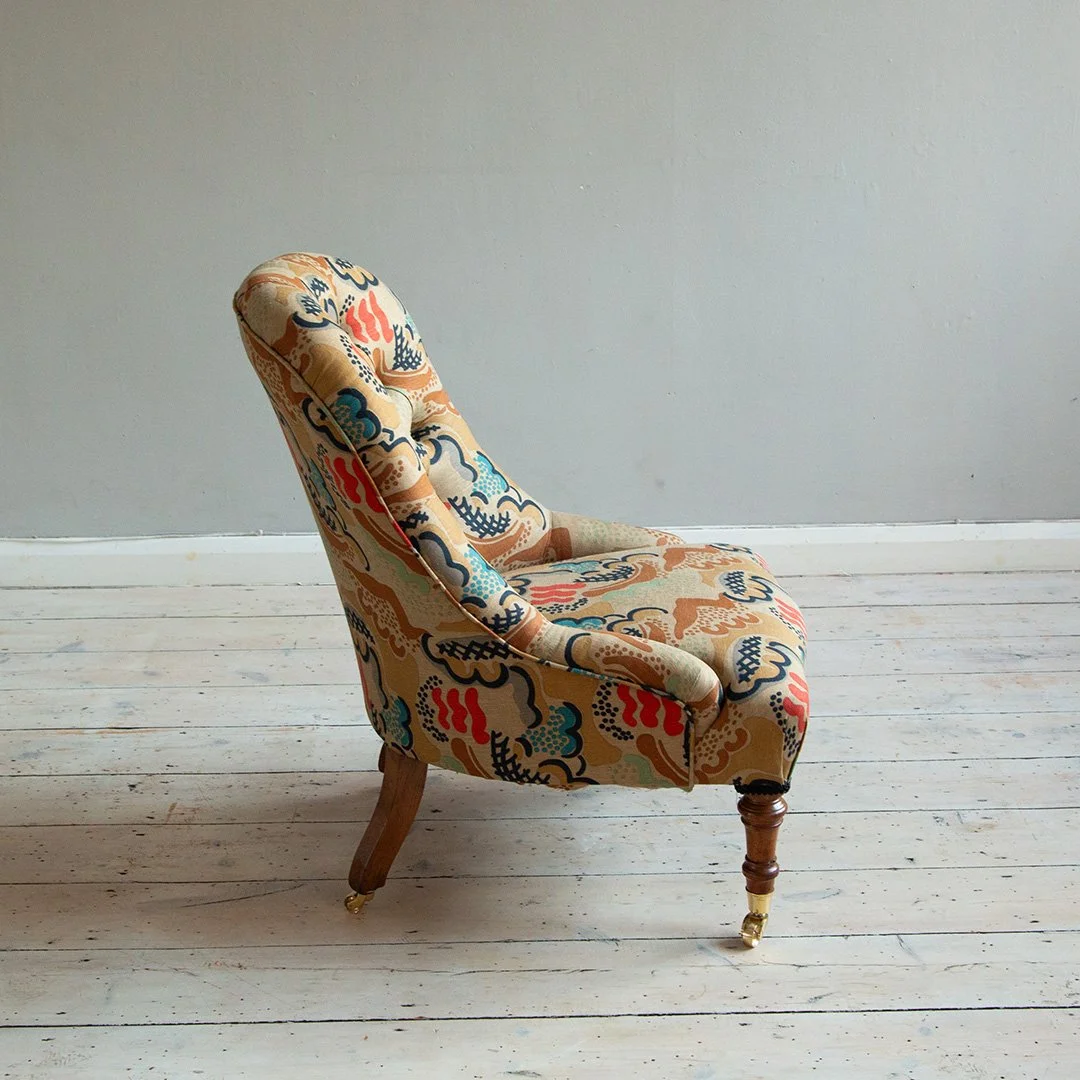 A vintage armchair with floral upholstery in beige, blue, red, black, and cream colors, standing on a light wood floor against a plain grey wall.